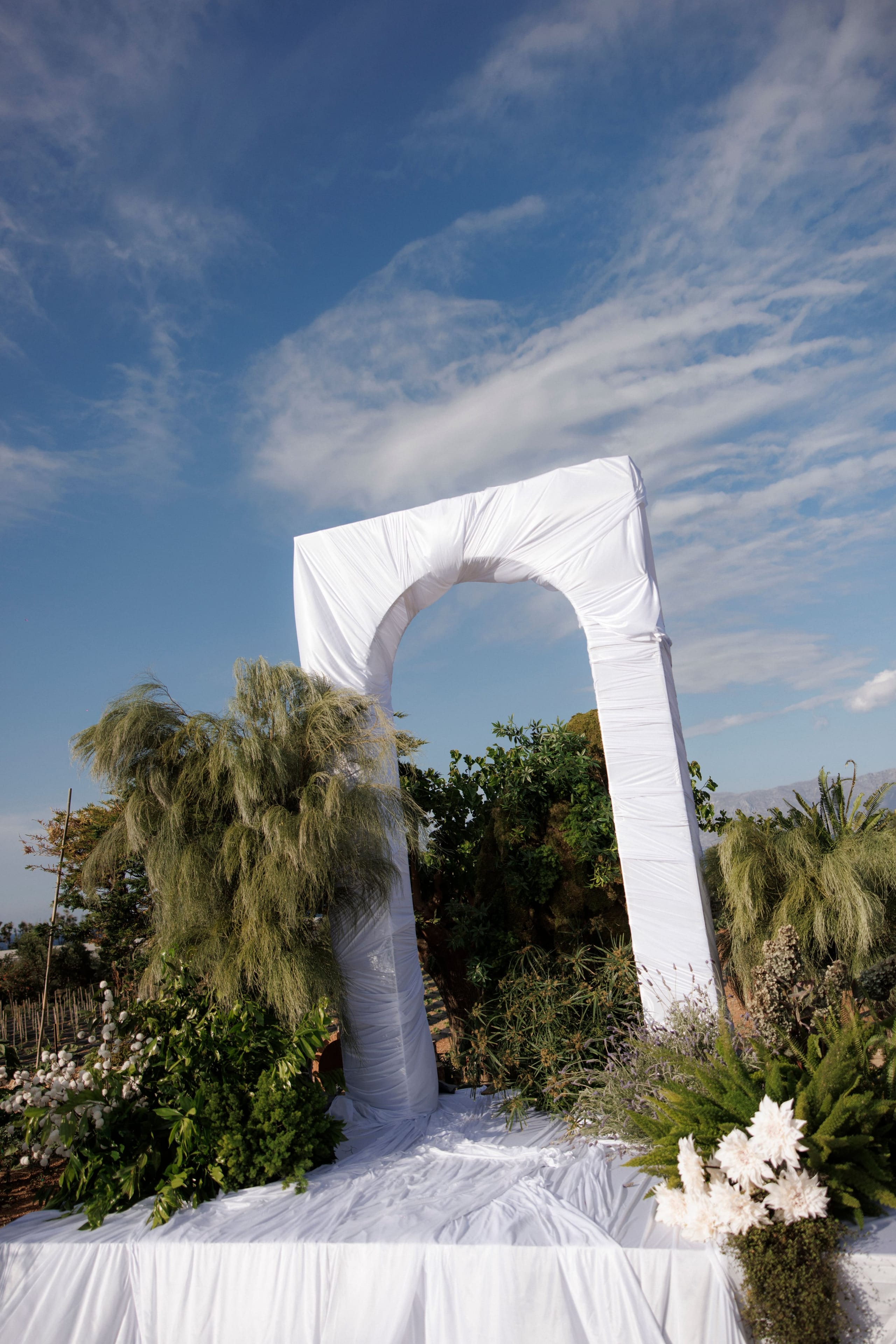 A tall white draped arch stands on a white-covered platform, framed by feathery grey-green grasses, leafy shrubs and clusters of white chrysanthemum-style blooms at its base, all set against a bright blue sky with wispy clouds. This wedding ceremony arch at Gramboussa Restaurant in Kaliviani, Crete, Greece, is dressed in soft folds of fabric and has a calm, airy, coastal feel.