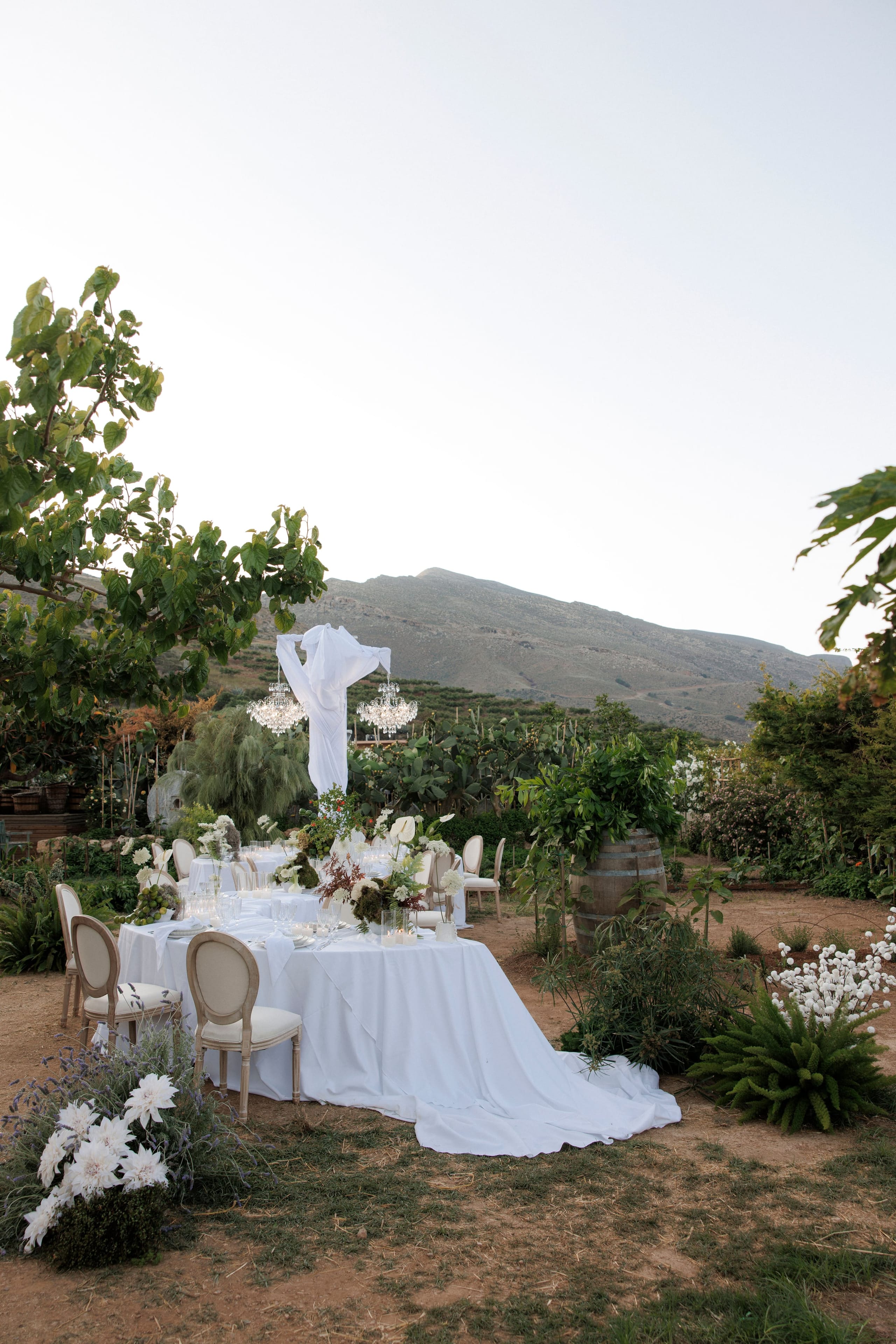 A long wedding table in an open garden at Gramboussa Restaurant in Kaliviani, Crete, Greece, dressed in a flowing white cloth that spills across the dry earth. Its low centrepieces mix white blooms with deep burgundy foliage and small candles, and round cream-upholstered chairs line the sides. Above it, a sheet of white fabric drapes from a pole between two crystal chandeliers, with a mountain ridge and soft pale sky behind, and a large white flower arrangement sits at the table's edge.
