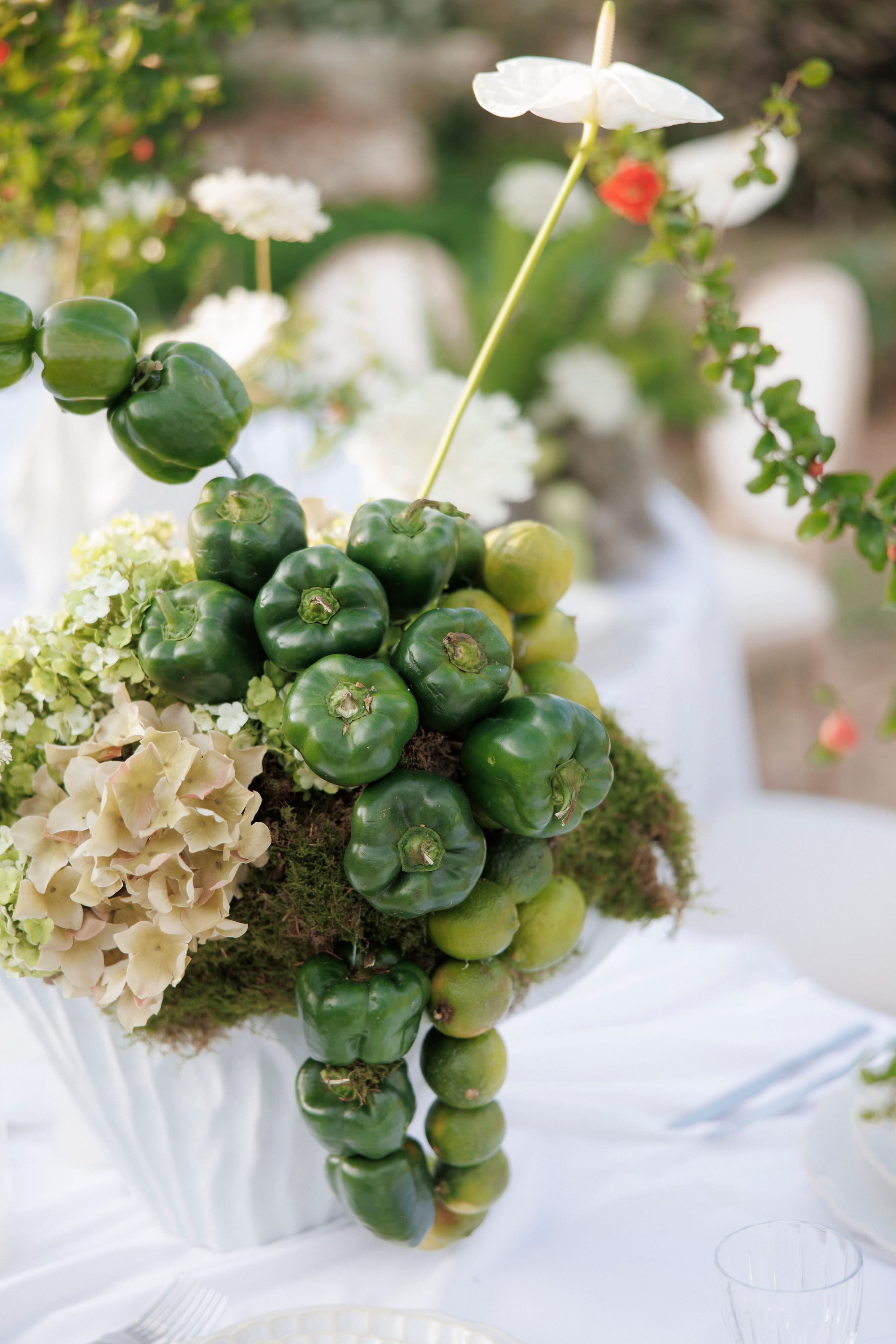 A white ceramic urn centrepiece on a white tablecloth, overflowing with glossy green bell peppers and small green limes cascading down its side, with moss at the base, pale cream hydrangeas, and a single white anthurium rising above. These wedding table flowers for Sara & Smith at Gramboussa Restaurant in Kaliviani, Crete, Greece, have a soft garden setting blurred behind them.