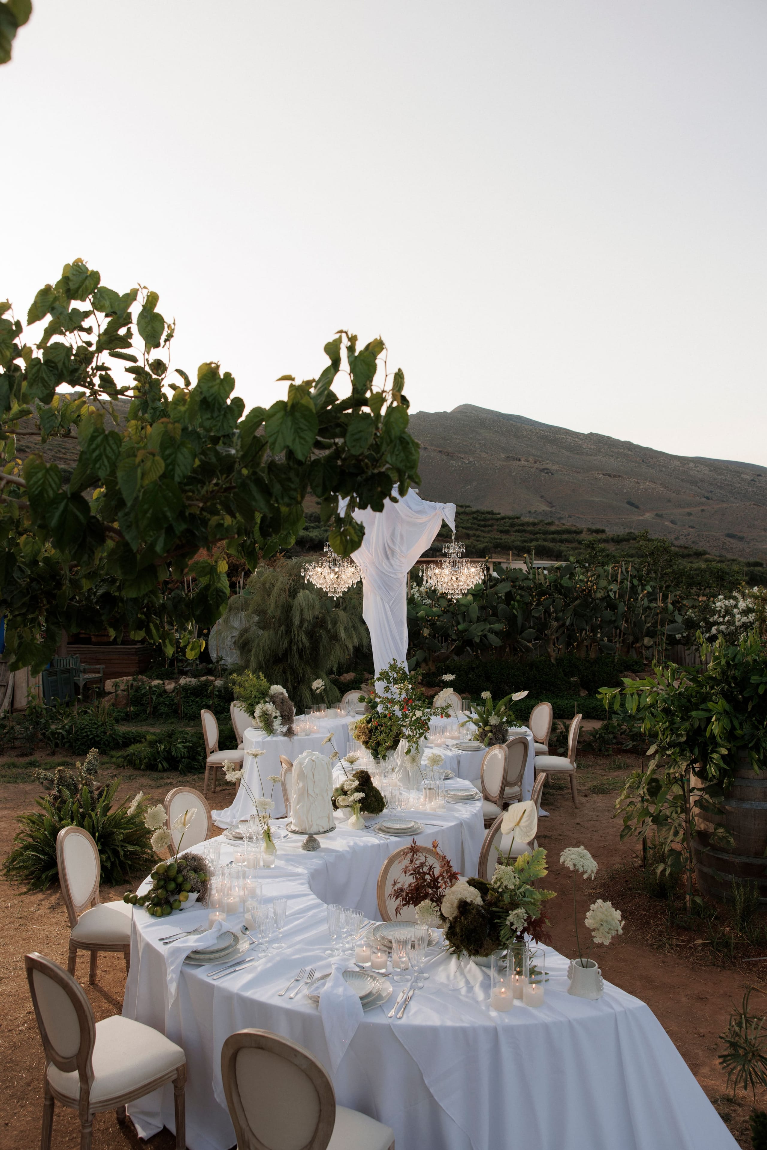 Long wedding tables draped in white linen wind across a dusty garden at Gramboussa Restaurant in Kaliviani, Crete, Greece, set with candles, clear glassware and low arrangements of white hydrangea and garden roses in soft green and bronze foliage. Crystal chandeliers hang from a fabric canopy in the background, with a rugged brown hill and fig-like leaves framing the warm evening sky. Cream cushioned chairs line the tables, creating a calm, romantic dinner setting for a wedding.