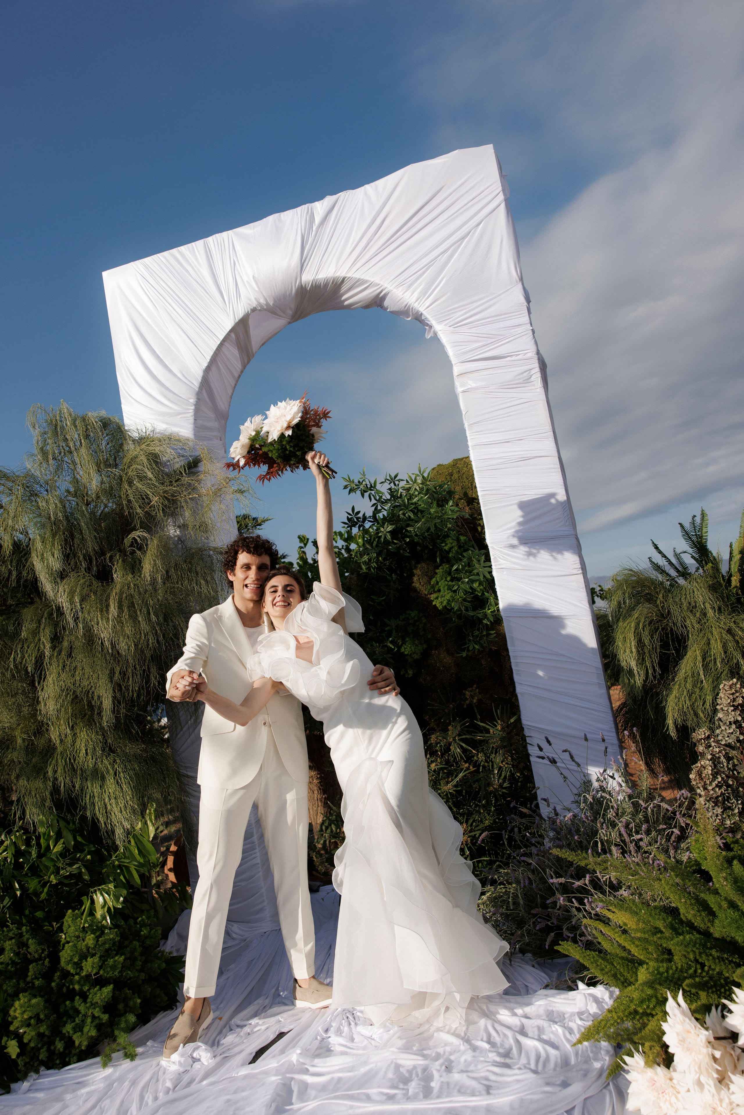 A bride in a flowing white gown with ruffled sleeves and her groom in a cream suit pose under a draped white arch in a garden at Gramboussa Restaurant in Kaliviani, Crete, Greece. She raises a bridal bouquet of white dahlias and rust-toned foliage high above her head while he holds her other hand and smiles beside her. Tall feathery green plants and white flowers frame them against a bright blue sky with wispy clouds.