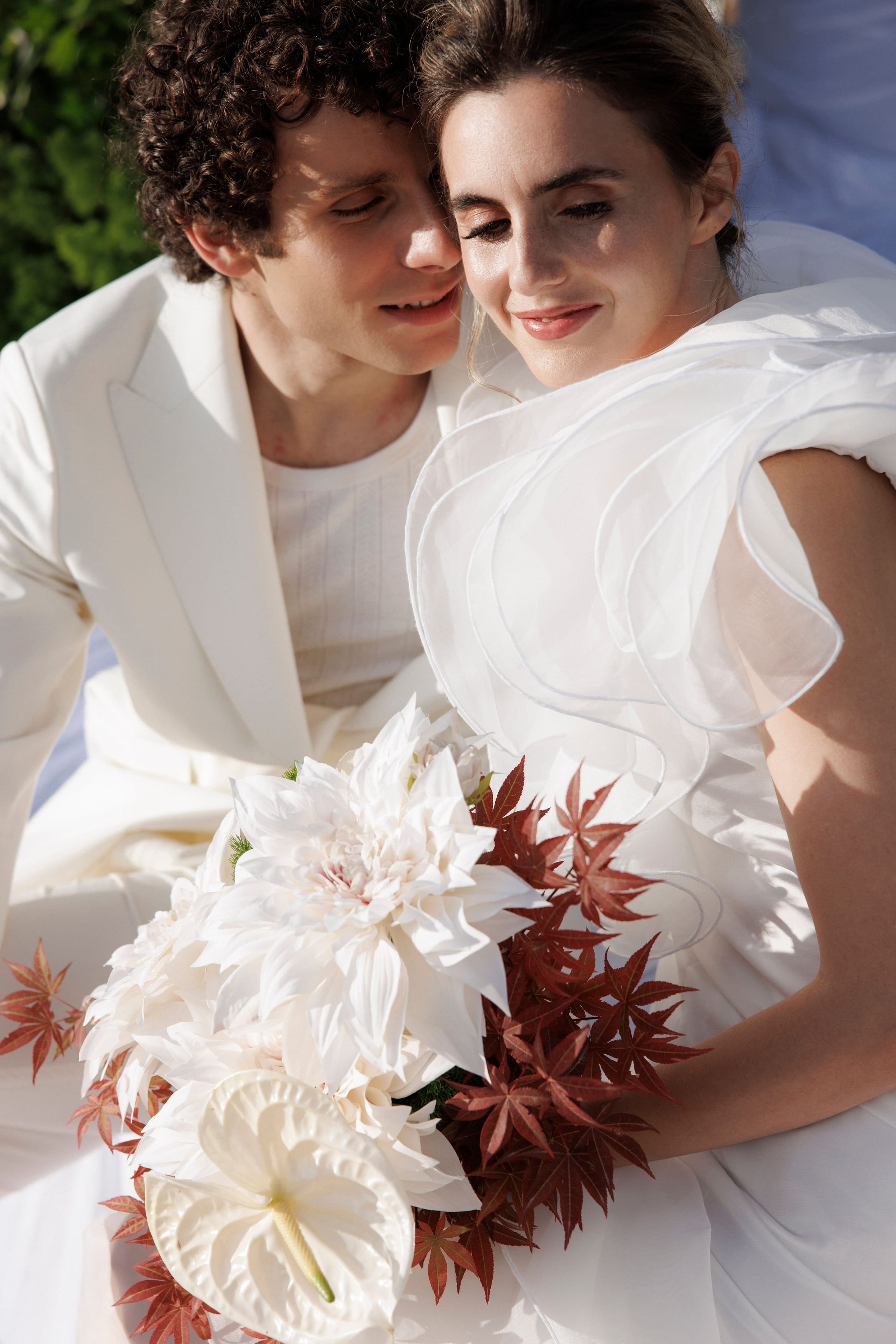 A bride and groom in white sit close together, their heads leaning in, both smiling with eyes lowered in the warm sunlight. She holds a bridal bouquet of cream dahlias and white anthurium, threaded with trailing burgundy Japanese maple leaves, while he wears an ivory suit and she wears a white gown with sculpted sheer ruffles on the shoulder. This wedding bouquet is from Greece, styled for a celebration at Gramboussa Restaurant in Kaliviani, Crete.