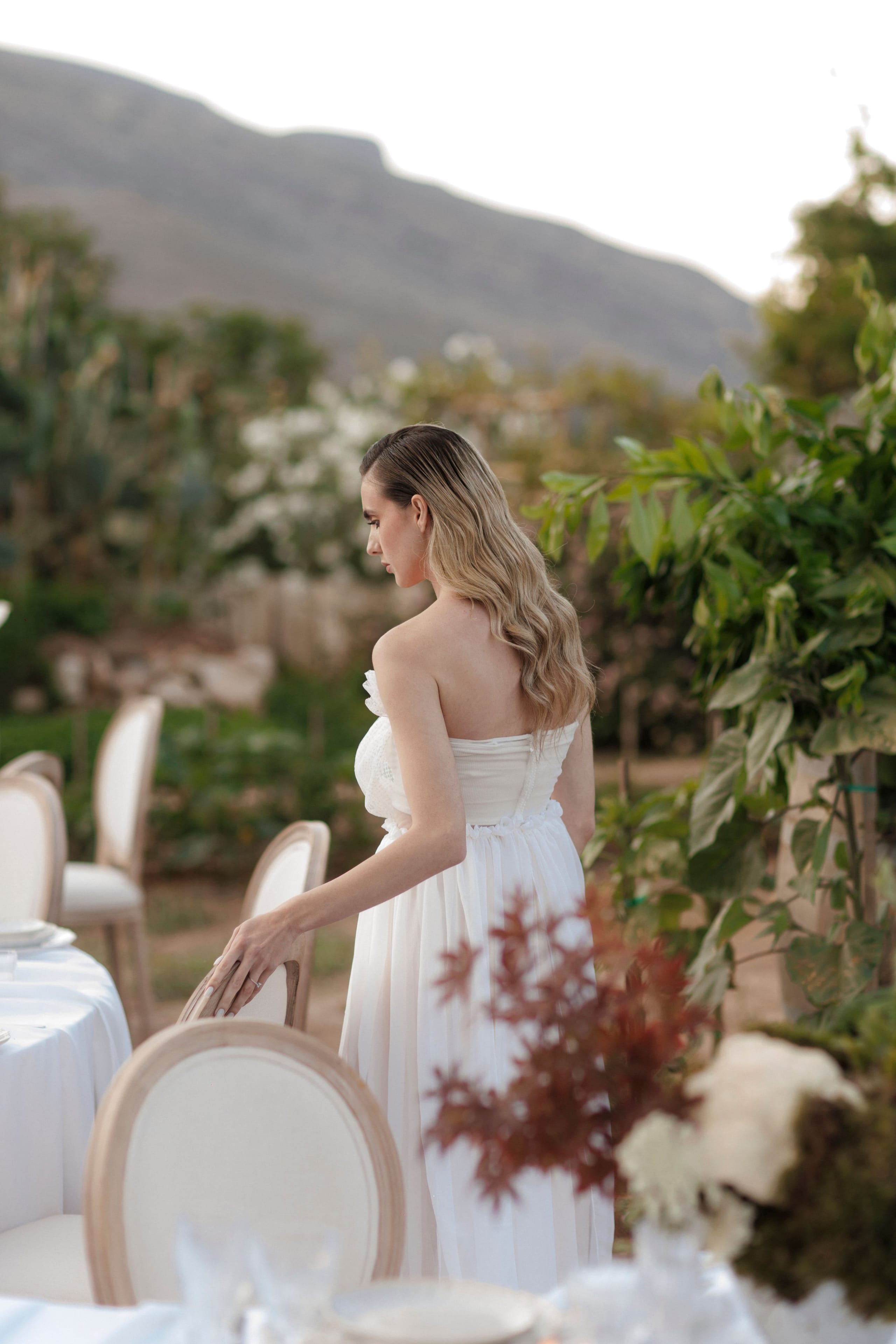 A woman with long, wavy honey-blonde hair stands with her back to us, one hand resting on the back of a cream upholstered chair beside a table dressed in white linen. She wears a flowing white strapless gown with a soft ruffled waist, and she gazes toward a green garden and a hazy mountain ridge. In the foreground, a rust-red Japanese maple sprig and creamy white blooms make up a wedding table centrepiece, at Gramboussa Restaurant in Kaliviani, Crete, Greece.