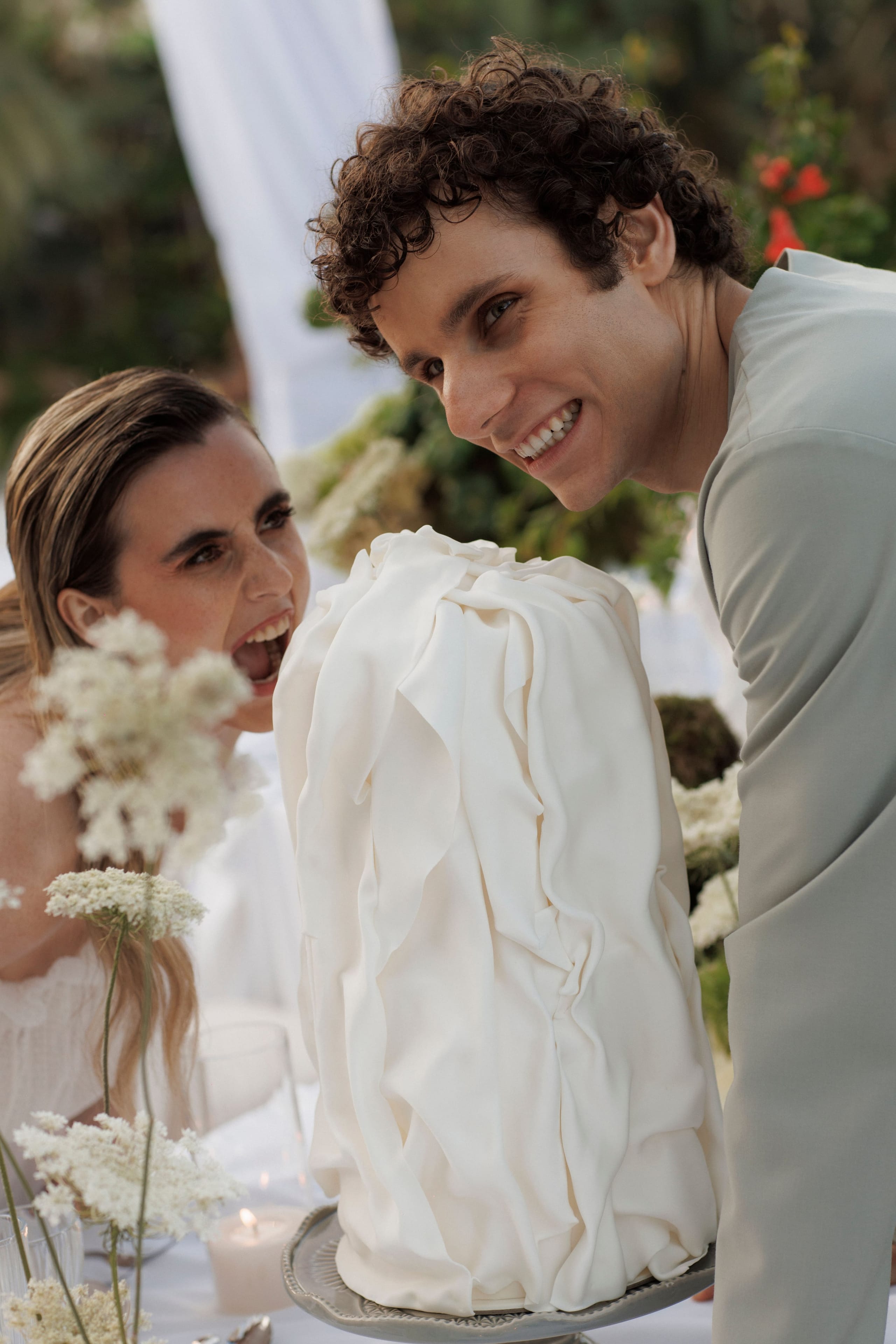 A man with curly dark hair and a sage-green suit leans in beside a bride with sleek, pulled-back hair, both smiling as she bites into a tall white wedding cake with soft, ruffled fondant folds. In the foreground, delicate white wild-carrot blooms and a small candle sit on a table at a wedding reception at Gramboussa Restaurant in Kaliviani, Crete, Greece, with greenery and white draping softly blurred behind them.