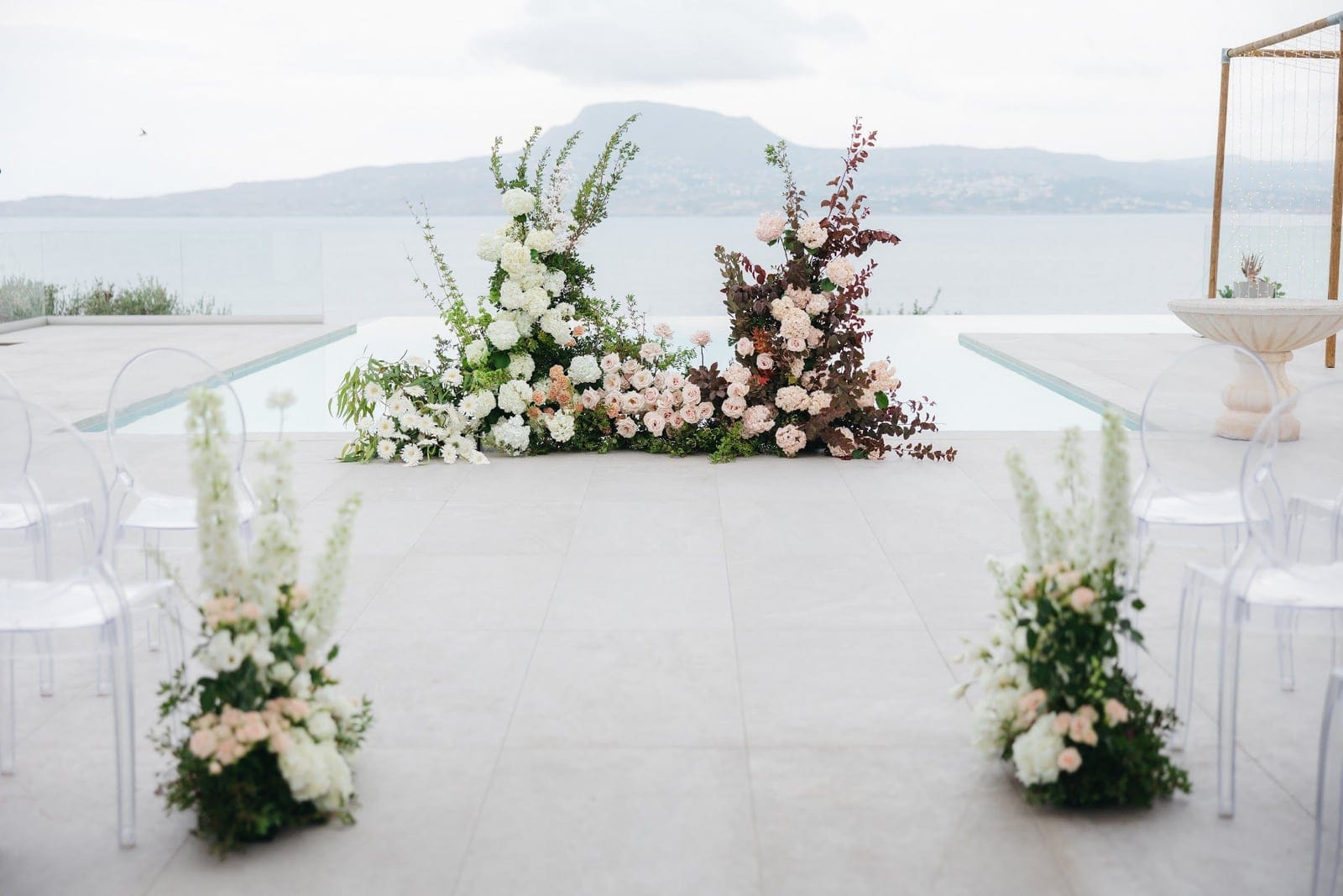 Wedding ceremony aisle at Villa Imperial in Crete, Greece, with clear acrylic chairs lining a pale stone terrace. Two tall floral arrangements of white hydrangeas, blush roses and dusky burgundy foliage frame a reflecting pool, with white delphinium spires and soft pink roses at the aisle edges. Behind them the sea stretches calm and misty toward a hazy mountain ridge, giving the scene a soft, airy, romantic feel.