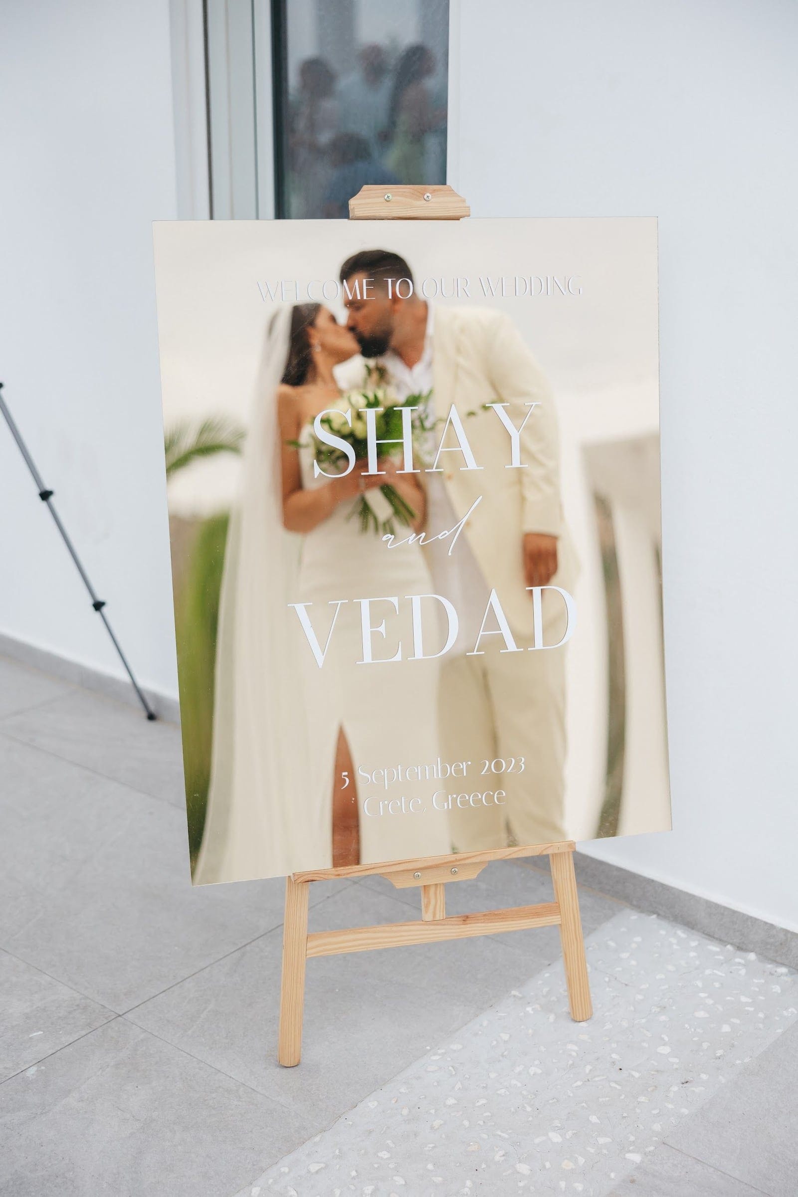 A mirrored welcome sign on a wooden easel reads "Welcome to our wedding" with "Shay and Vedad" in large white lettering, and through the glass a blurred photograph of a bride in a fitted white gown and long veil kissing her groom in a cream suit, holding a loose bouquet of white roses and green foliage. This wedding sign is at Villa Imperial in Greece, set against a plain white wall.