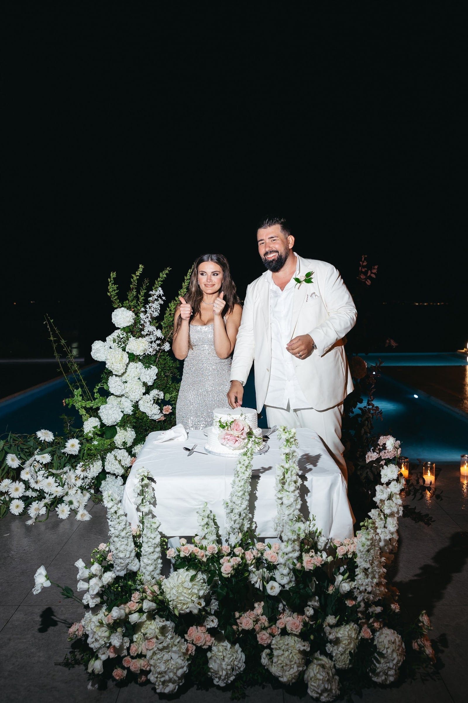 A bride in a sparkling silver sequined gown and a groom in a white suit stand smiling at a white-draped cake table at night, both giving a thumbs up, at a wedding in Greece. The table is surrounded by white hydrangeas, white stock, white daisies and blush garden roses, with dark foliage spilling down the sides, and a pool glows blue behind them with candles flickering at the edge. This wedding flower display is at Villa Imperial in Crete.