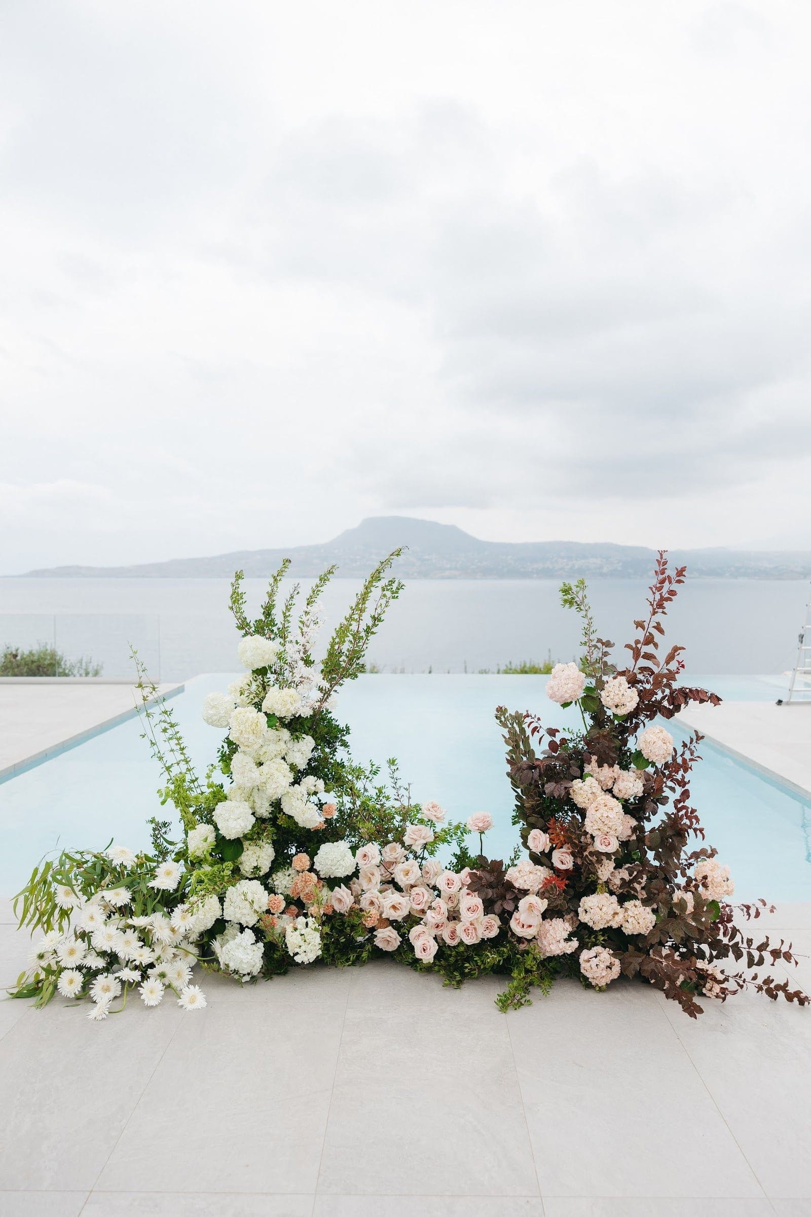 Three tall floral installations stand along the edge of an infinity pool at Villa Imperial in Crete, Greece, with the sea and a hazy mountain behind them under a soft grey sky. On the left, white hydrangeas, white daisies and green foliage rise in a full cascade, while the centre is a lush mix of blush garden roses, peach blooms and greenery. On the right, deep burgundy branches frame cream and blush hydrangeas and roses. These wedding flowers are a calm, airy palette of white, blush and plum.
