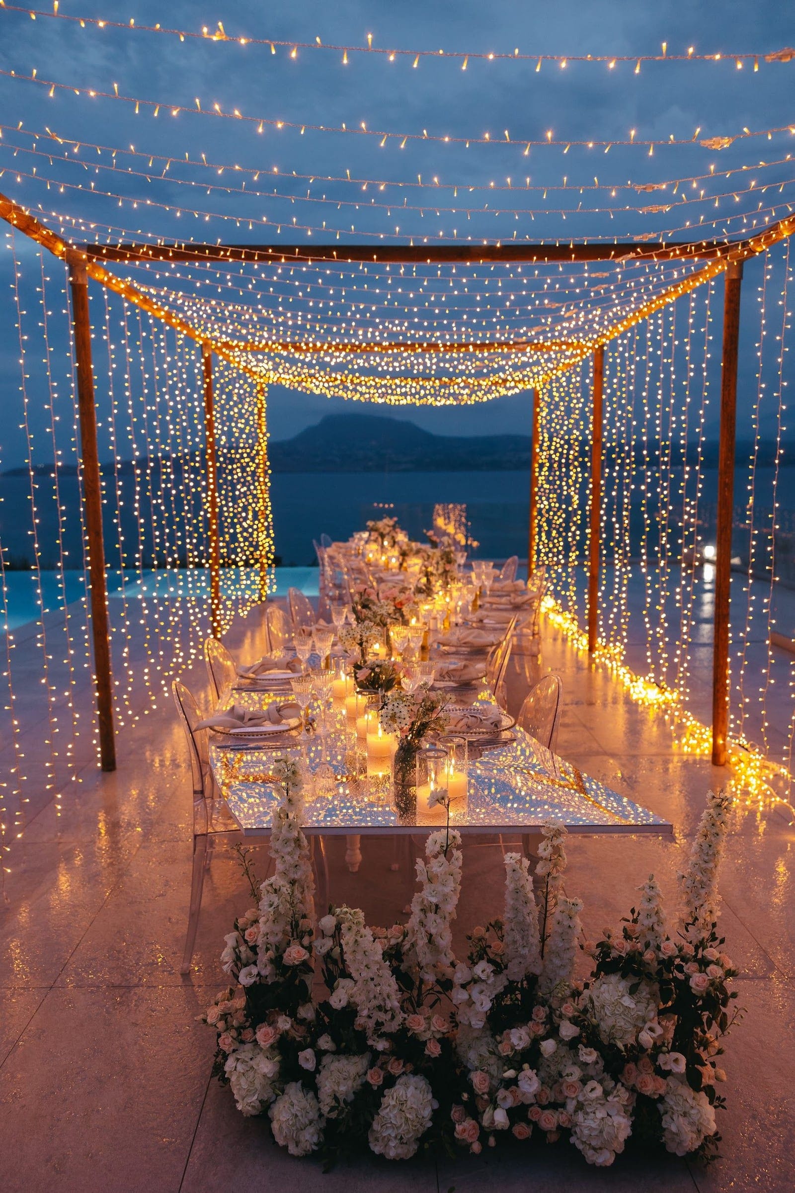 A long wedding dining table under a canopy of warm fairy lights, set with clear acrylic chairs, candles and white linen, at Villa Imperial in Crete, Greece, as dusk settles over the sea. Along the front of the table runs a dense low arrangement of white hydrangeas, delphinium spires and soft blush roses, and the lit cords drape down on either side like a glowing curtain. Beyond the open sides, a calm blue sea and a dark silhouetted hill frame the evening sky.