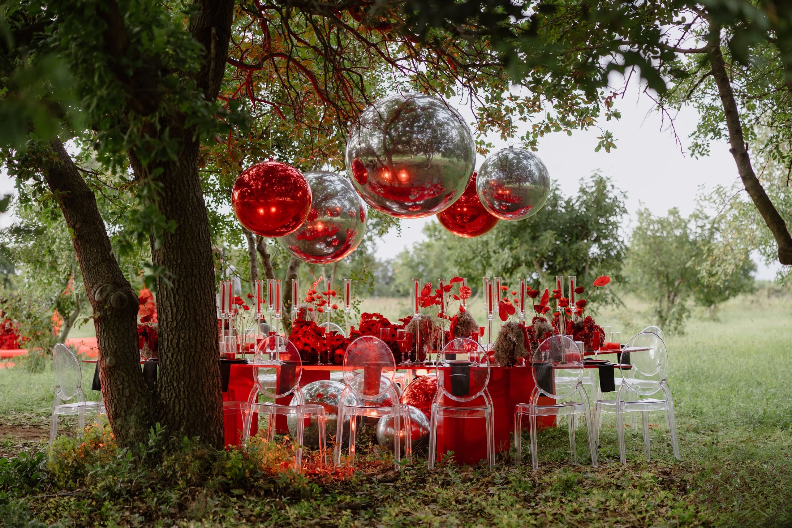 A long outdoor wedding table under an olive tree in Rethimno, Crete, Greece, dressed in red from cloth to centrepiece. Red and silver mirrored spheres hang from the branches above, reflecting the table and greenery, while clusters of red roses and poppies sit among tall taper candles. Clear ghost chairs line the table on the grass, and the soft overcast light gives the scene a dreamy, intimate feel.