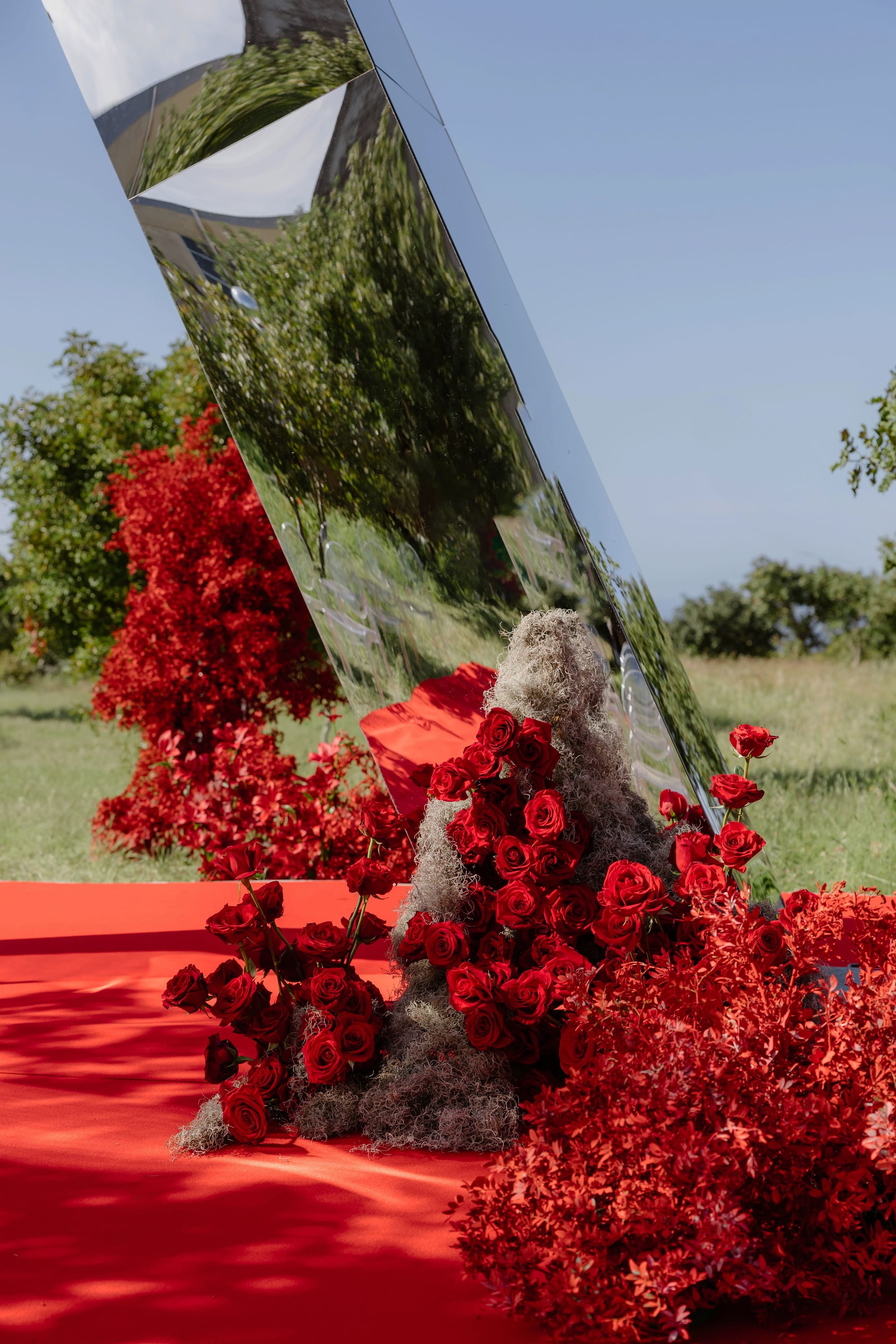 A tall, slanted mirrored panel leans over a mound of deep red roses cascading down a red carpet, with grey lichen-like moss tucked between the blooms and bright red foliage spilling out at the base, all set on a grassy lawn under a clear blue sky. This wedding flower installation for Silia and Louis, in Rethimno, Crete, Greece, reflects the surrounding trees and sky in the shining mirror surface.