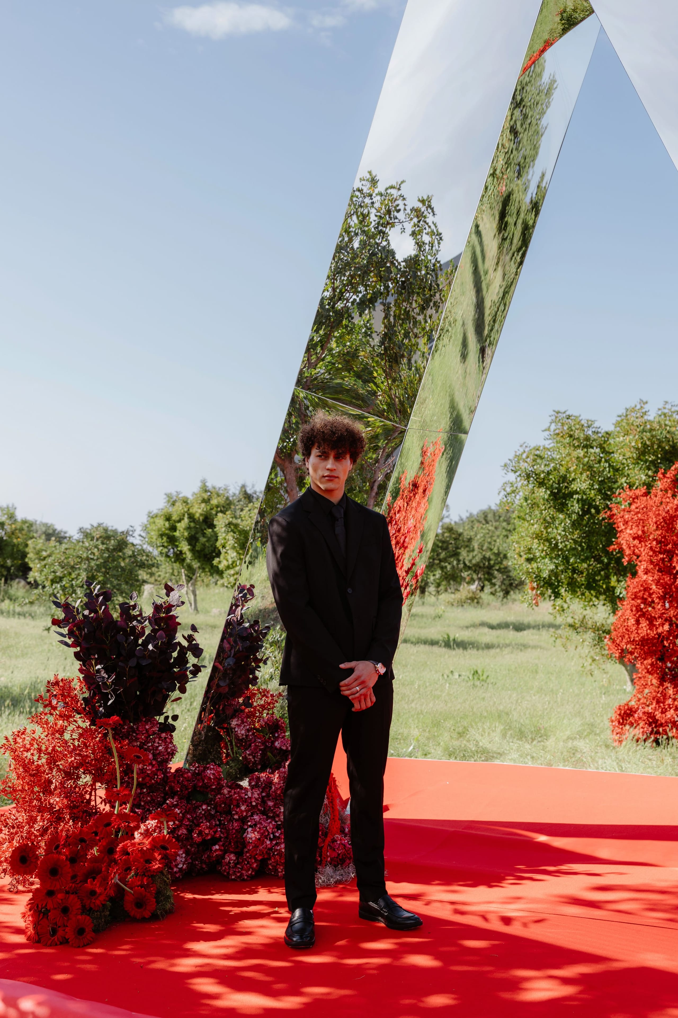 A young man with curly dark hair stands in a black suit and tie on a red carpet, hands folded in front of him, beside a tall angled mirror panel that reflects the trees and sky. At his feet sit clusters of deep red sunflowers, dark plum-coloured foliage and burgundy hydrangeas, with bright red blooms spilling from the shrubs at the right. This wedding flower display for Silia and Louis, in Rethimno, Crete, Greece, is set in a sunlit olive grove under a clear blue sky.