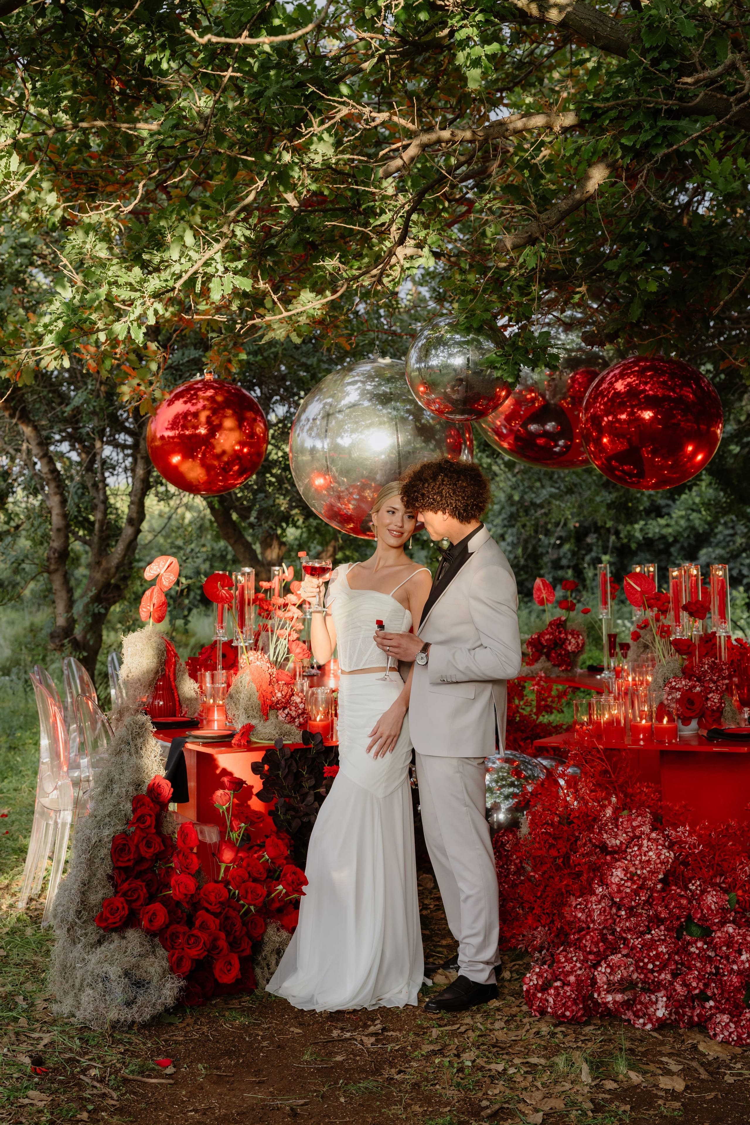 A woman in a white satin crop top and long white skirt holds a glass of red wine, leaning in beside a man in a light grey suit beneath a canopy of trees hung with large red and silver mirrored spheres. Along the red-draped table below, red roses, red anthurium, and clusters of red and white hydrangeas spill across the grass, with tall clear candle holders glowing among them. This wedding table flower styling is set outdoors in Rethimno, Crete, Greece.