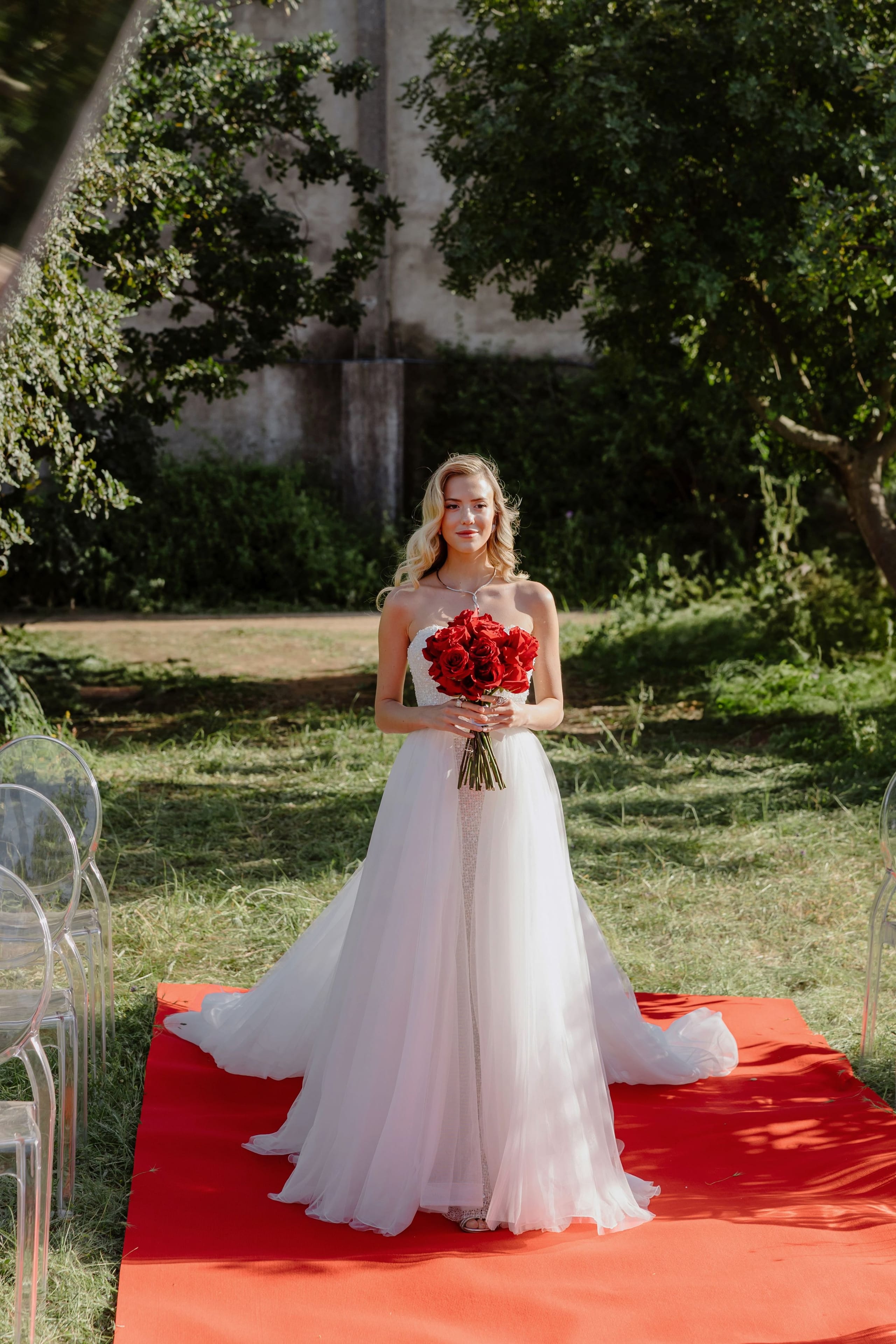 A bride with wavy blonde hair stands on a red aisle runner in a grassy garden at a wedding in Rethimno, Crete, Greece, holding a tight bouquet of deep red roses. Her strapless white tulle gown flows into a soft train, and she wears a delicate necklace. Clear acrylic chairs line the aisle beside her, and dappled sunlight falls through the trees behind her, creating a warm, romantic mood.