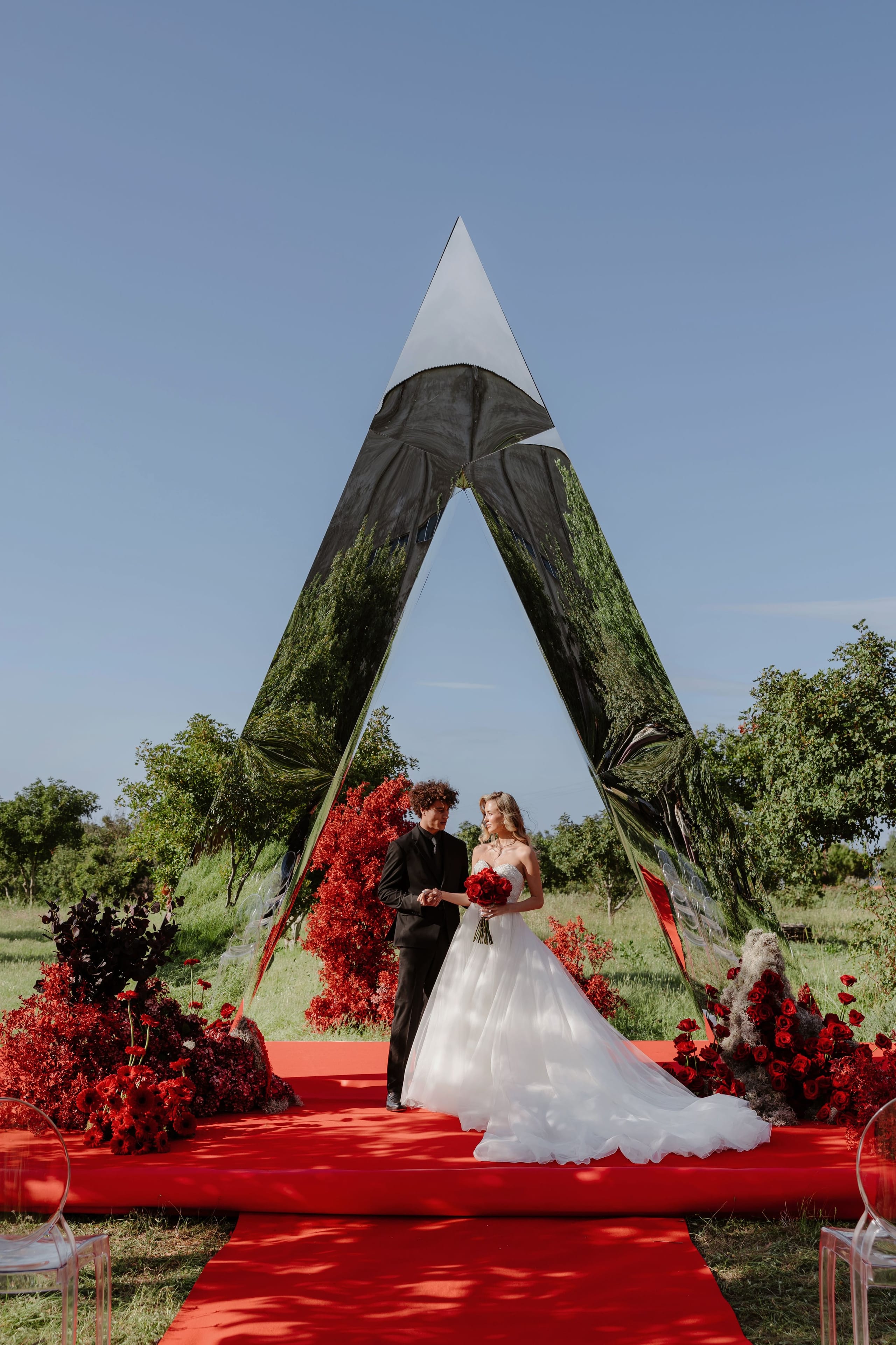 A bride in a strapless white ballgown with a flowing tulle train stands beside a groom in a black suit, holding hands on a red carpet beneath a tall, mirror-polished triangular arch in an olive grove in Rethimno, Crete, Greece. Lush arrangements of deep red roses, crimson foliage and a dark plum-toned bloom frame the arch, and the bride carries a matching red rose bouquet. The clear blue sky and soft afternoon light make this a bold, striking wedding ceremony setting.