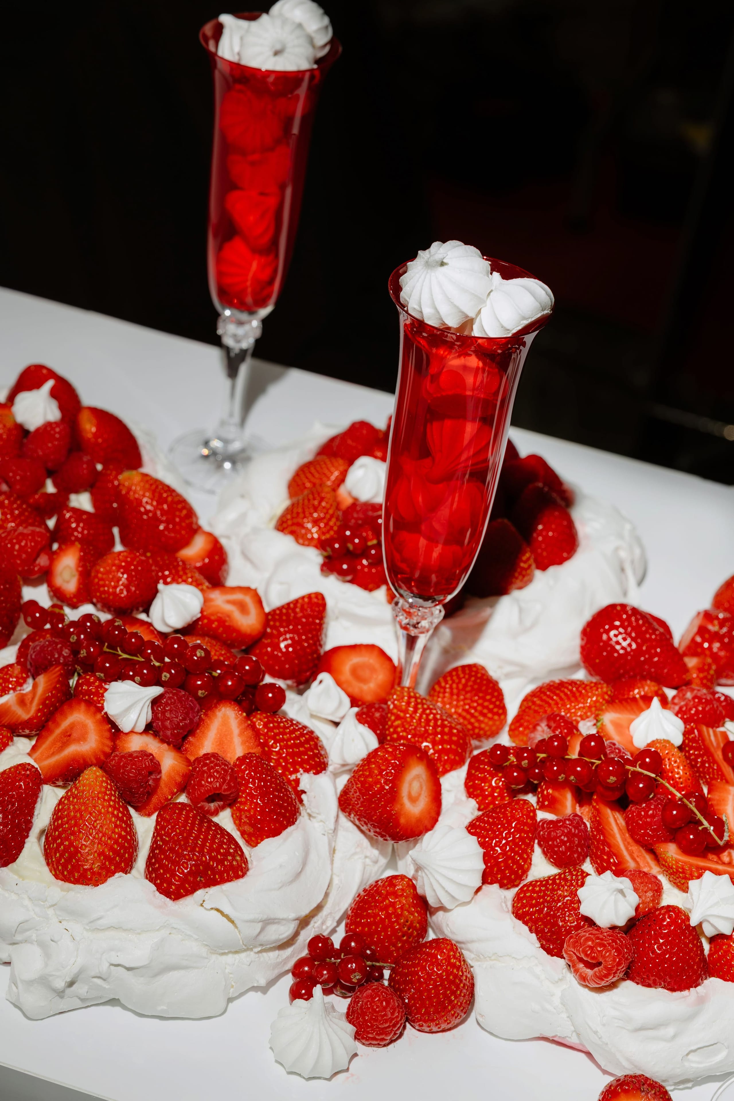 Tall champagne flutes filled with red strawberries and topped with meringue kisses stand among several white meringue pavlovas piled high with fresh strawberries, raspberries and redcurrants on a white table, a dessert display for a wedding at a venue in Rethimno, Crete, Greece. The dark background makes the glowing reds and creamy whites feel vivid and celebratory.