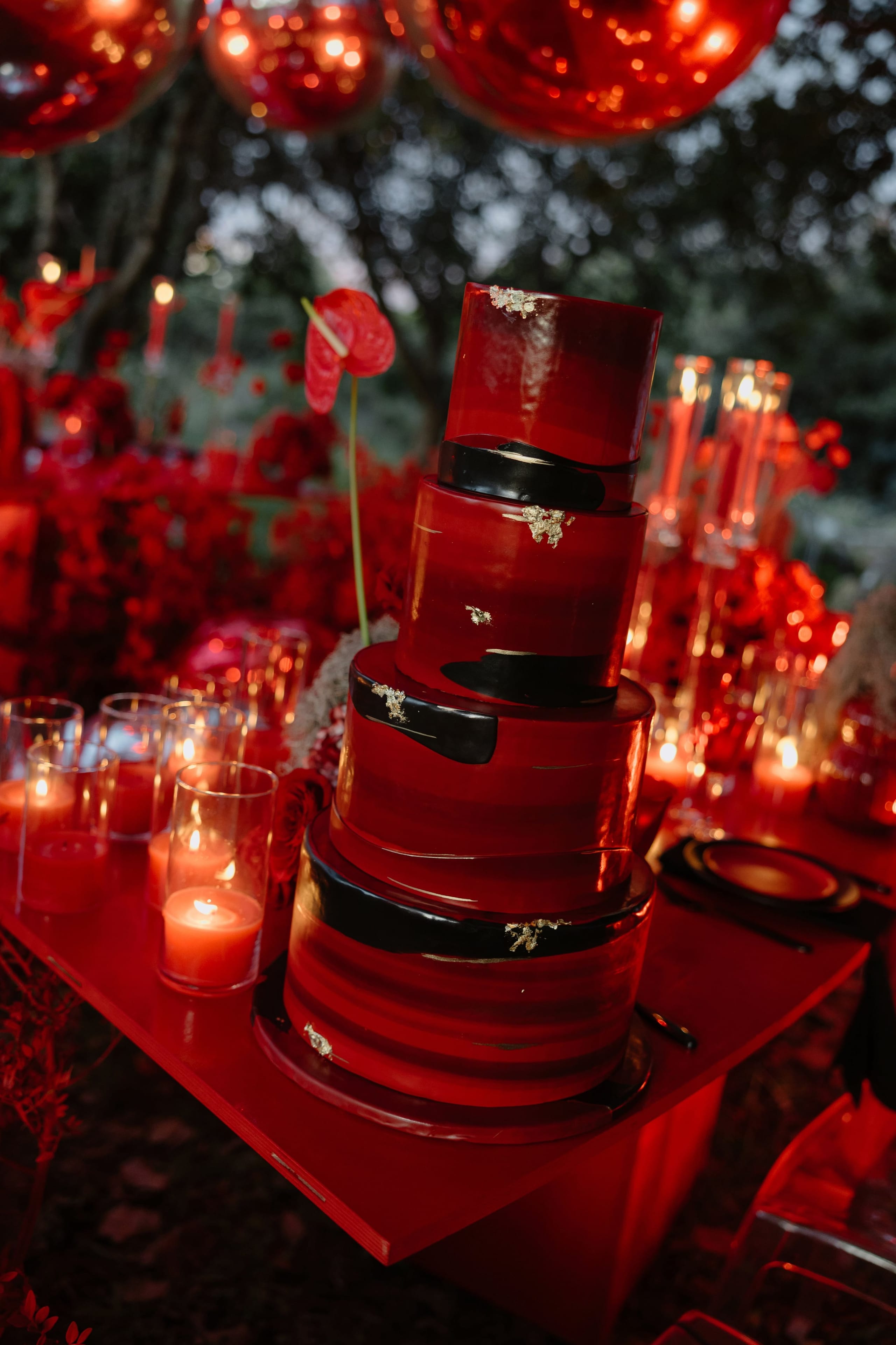 A tall four-tiered wedding cake in deep glossy red, banded with black ribbons and flecked with gold leaf, stands on a red table at an outdoor wedding in Rethimno, Crete, Greece. Behind it, a red anthurium blooms on a slender stem, with clusters of red roses, flickering glass votive candles and dangling red mirrored balls overhead, all set against dark trees in the evening light.