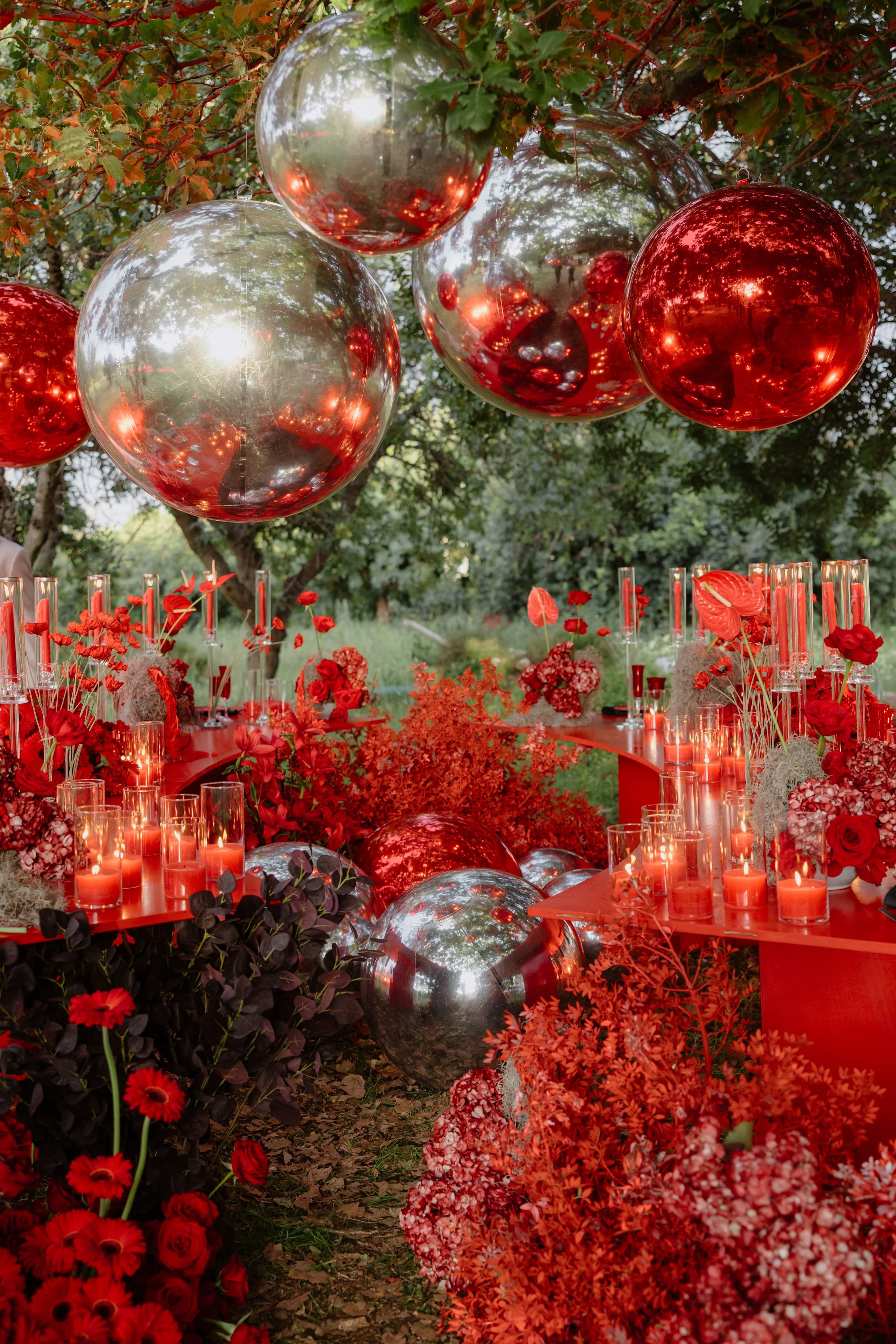 Hanging from the branches overhead are large mirrored spheres, alternating silver and red, above a long curved red table in an open garden. The table is set with glowing pillar candles in glass holders, red anthuriums, red gerberas and deep red roses, with pink and red hydrangeas and sprays of russet foliage spilling onto the grass. This is a wedding table styled with flowers in Rethimno, Crete, Greece, and the whole scene feels dramatic and celebratory in the evening light.