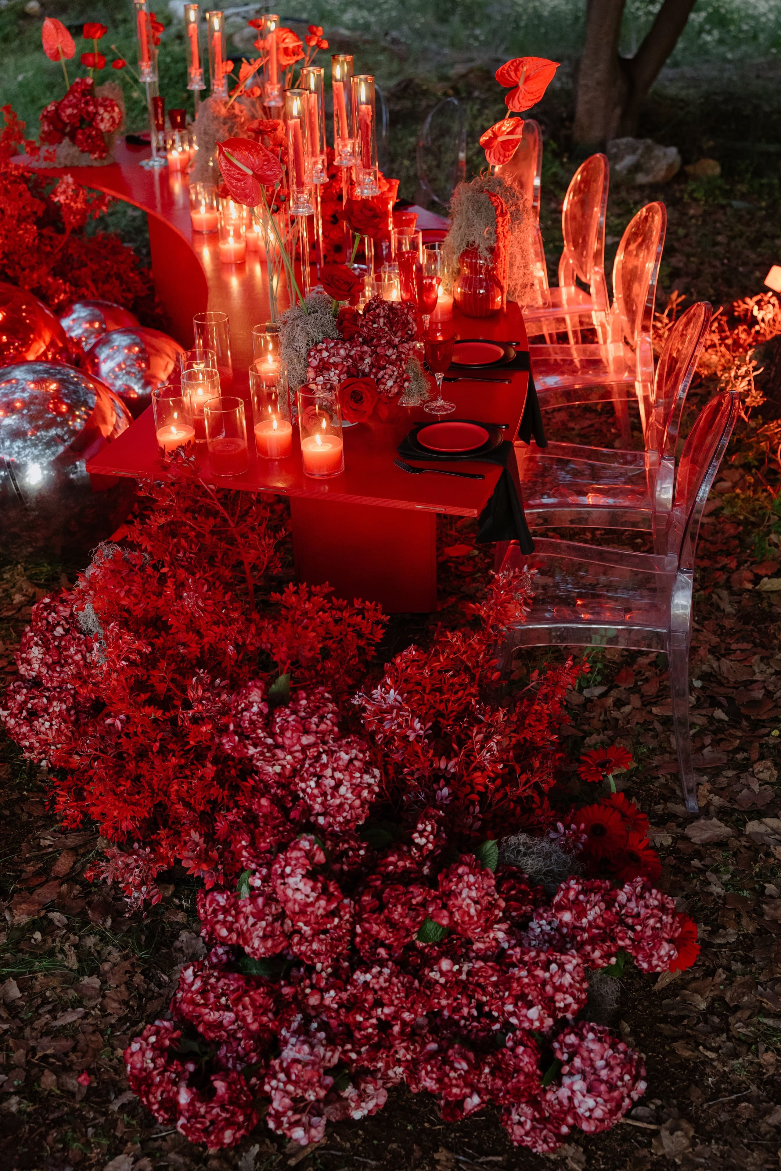 A red-themed outdoor wedding table in Rethimno, Crete, Greece, glowing in warm evening light. Red anthurium blooms rise among tall glass candle holders with pillar candles, and the low red table is framed by drifts of red and white speckled hydrangea and red foliage spilling across the ground. Clear acrylic chairs and red and black place settings complete the dramatic, romantic scene.