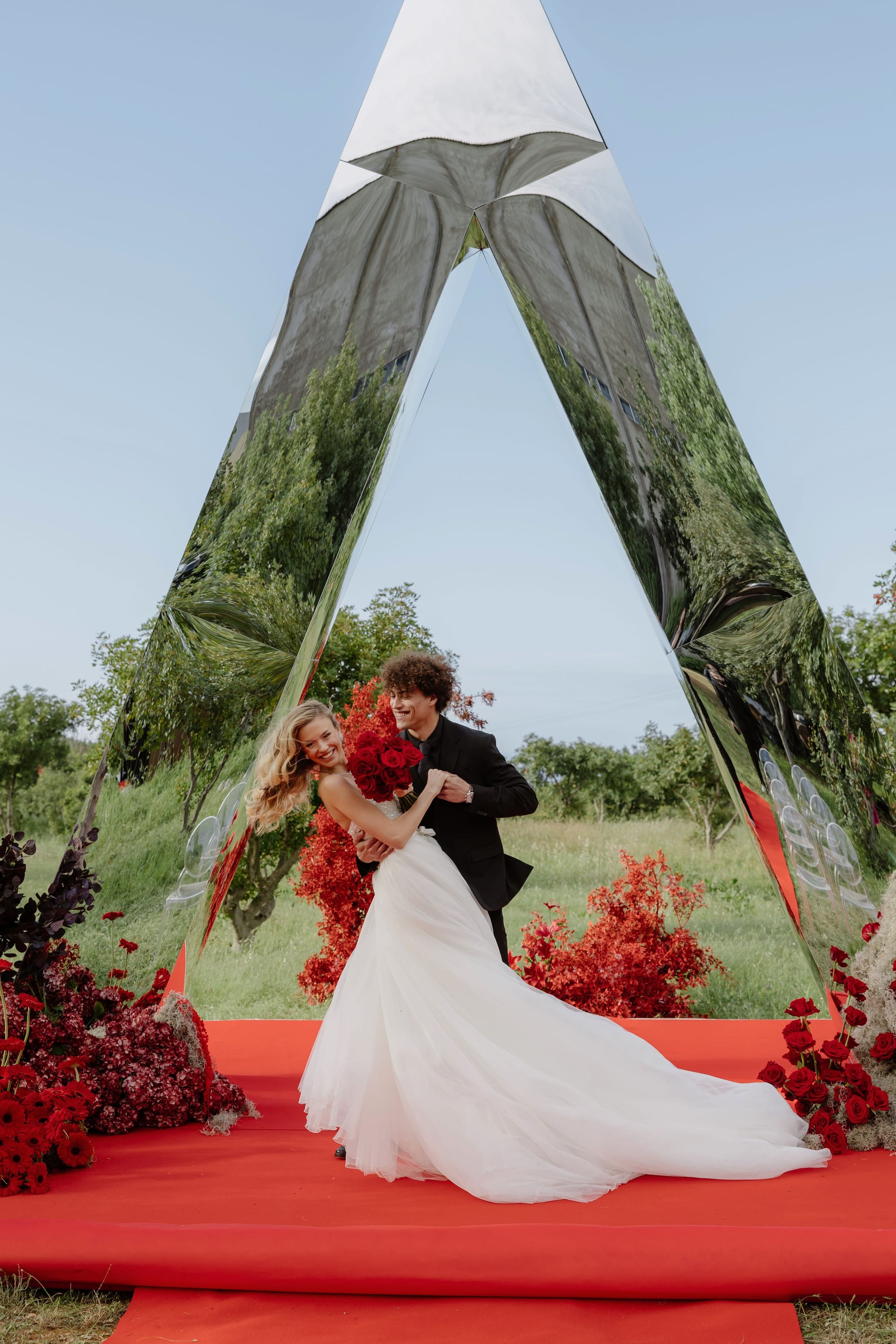 A bride in a flowing white gown laughs as a dark-suited groom lifts her slightly, both holding a bouquet of deep red roses, inside a tall triangular mirrored arch on a red carpet. Around them are red gerbera daisies, dark burgundy hydrangeas, red roses and red-leaved shrubs, with orchard trees reflected in the mirrors. This wedding flower design is for a wedding at Rethimno in Greece.