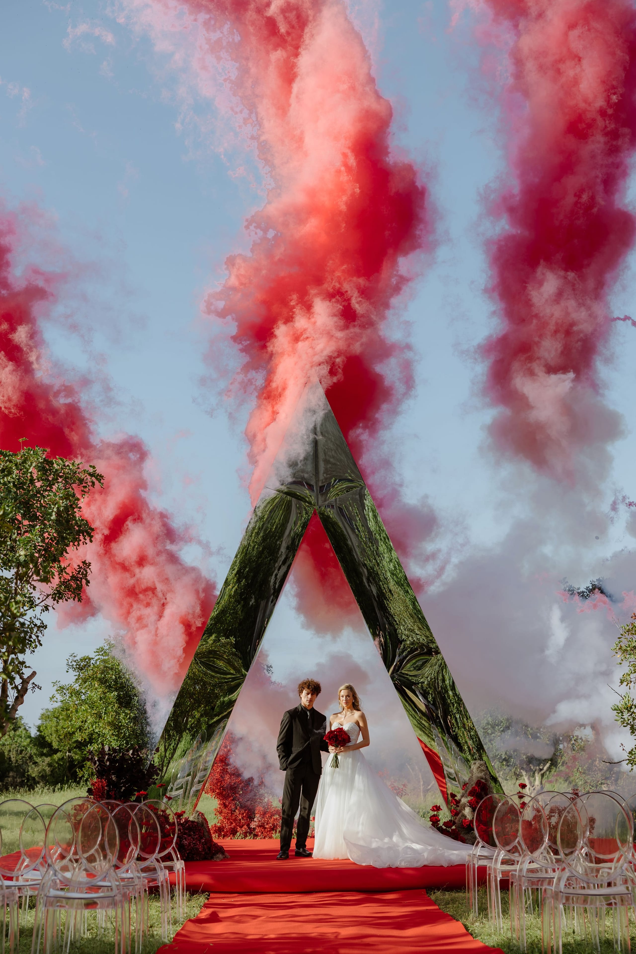A bride in a strapless white gown with a long train and a groom in a black suit stand side by side under a tall, triangular mirrored arch on a red-carpeted aisle, the bride holding a bouquet of deep red roses. Thick plumes of red and pink smoke rise behind the arch against a clear blue sky, while clear acrylic chairs line the aisle and dark red blooms frame the ceremony. This wedding flower display is set in Rethimno, Crete, Greece.