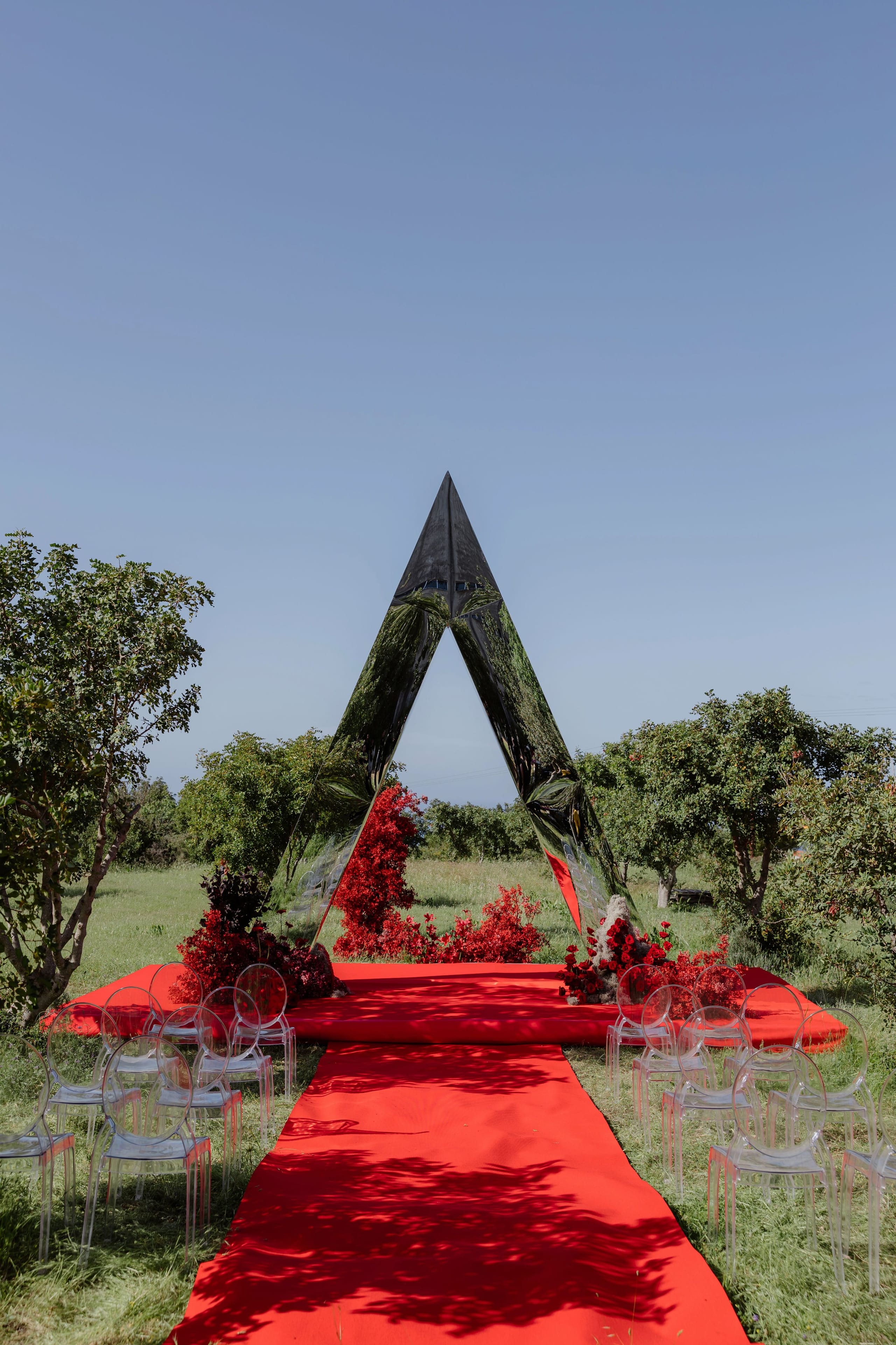 A tall, dark triangular arch stands in an open grassy orchard, framing a clear blue sky, with a red carpet running down the centre aisle toward it. Red and burgundy blooms, likely roses and bougainvillea-style clusters, are arranged around the base of the arch, with clear acrylic chairs lining both sides of the aisle. This is a wedding ceremony set-up in Rethimno, Crete, Greece, with a bold, modern look built around the vivid red and the dark structure.