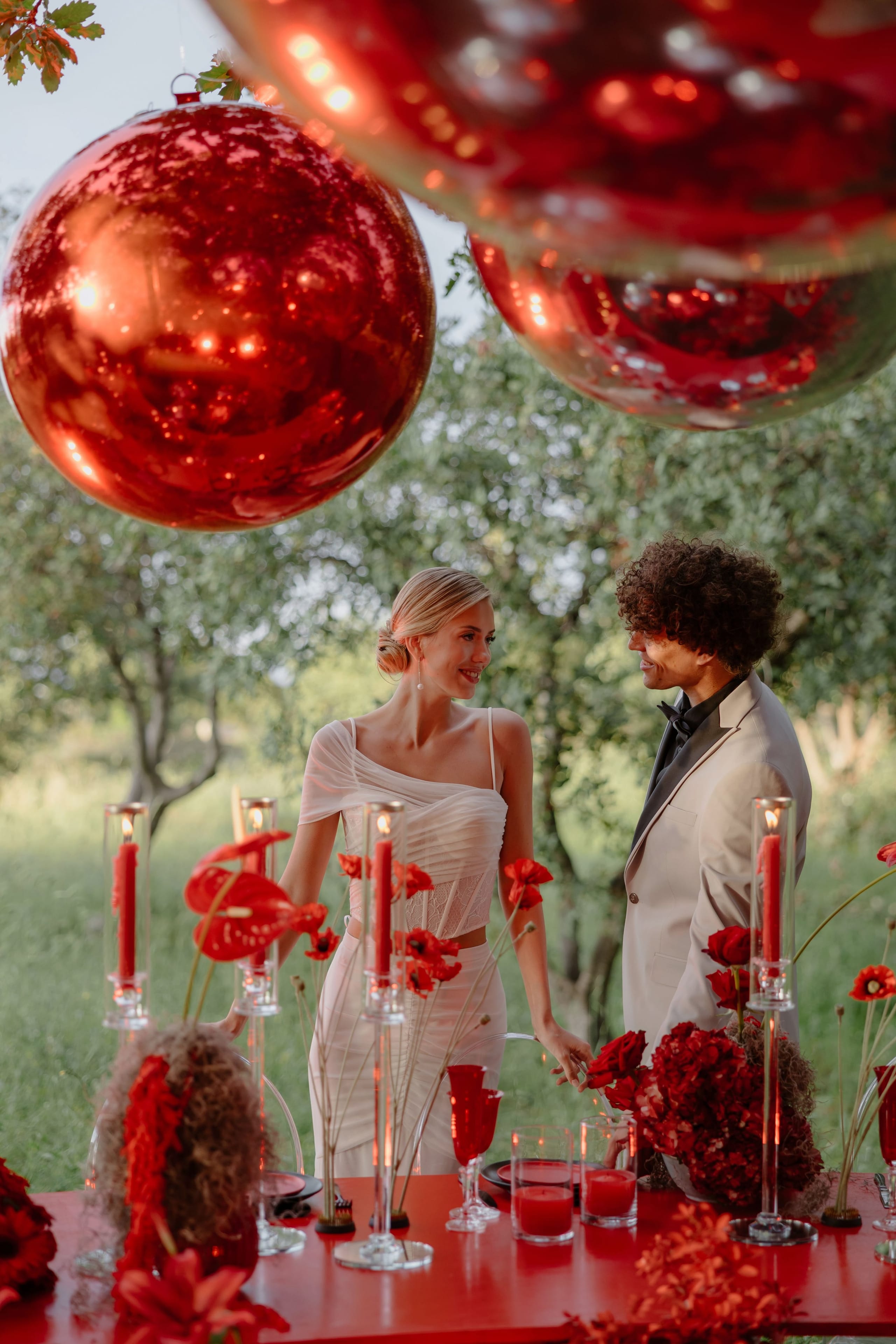 A bride in a pale, sheer-layered corset top and a groom in a cream jacket with a dark shirt stand smiling at each other behind a long red-topped table in an olive grove at Rethimno, Crete, Greece. Above them hang oversized glossy red baubles, and the table is dressed with red anthurium, red roses and hydrangeas, tall glass candle holders with red candles, and red glasses. This wedding table styling is set against soft green trees in gentle evening light.