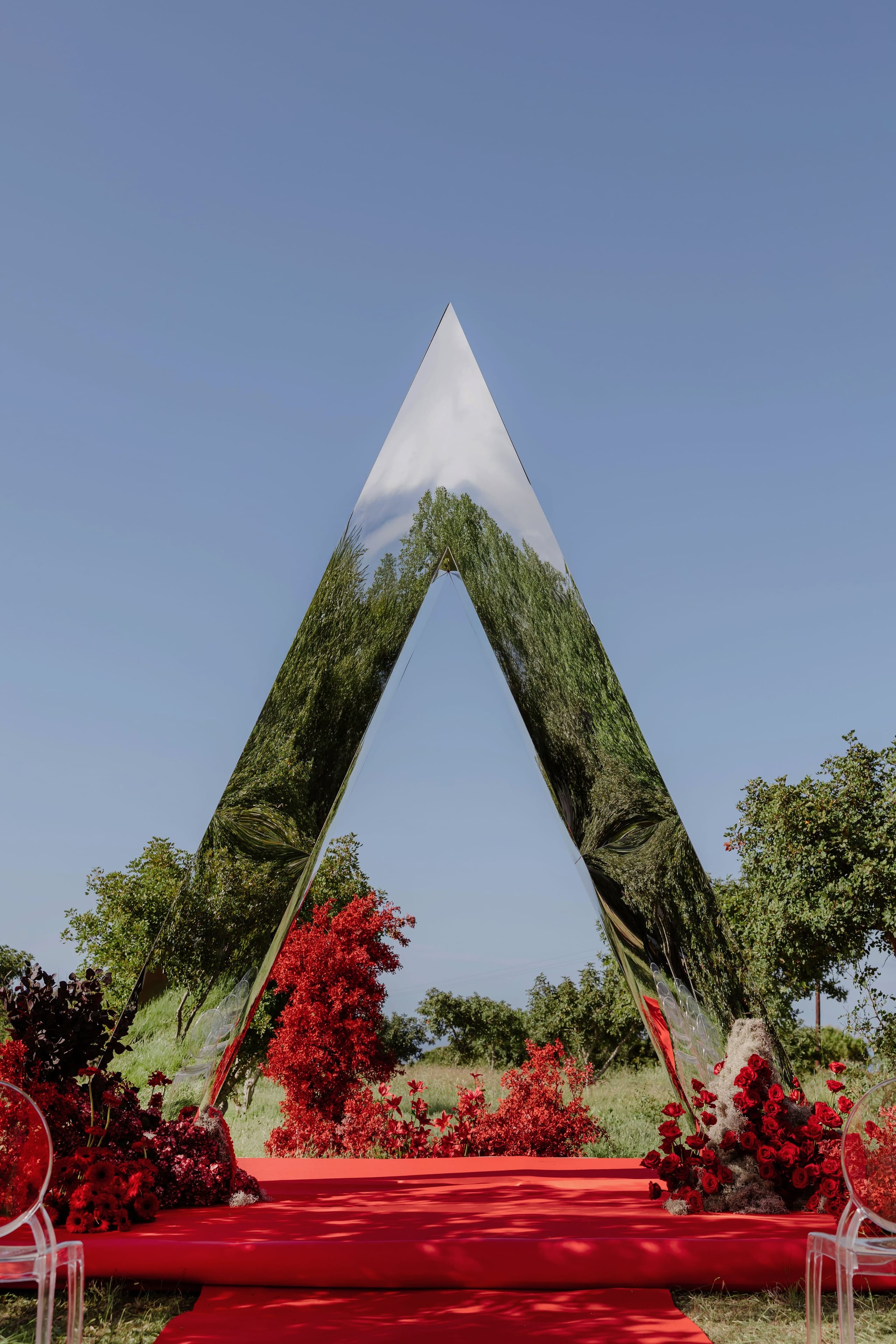 A tall, pointed arch of mirrored metal rises against a clear blue sky, reflecting the green trees and grass around it. At its base sit clusters of deep red roses, crimson foliage and red shrubs beside a red aisle runway, with clear acrylic chairs at the edges. This wedding ceremony arch, set in a grassy field for a wedding in Rethimno, Crete, Greece, is bold and striking in the bright summer light.