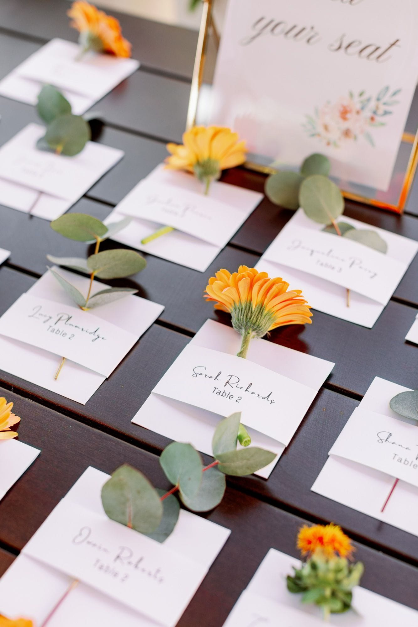 A close-up of place cards laid out in rows on a dark wooden table, each card tucked around a stem with a single orange calendula or gerbera-style bloom and a sprig of silver-green eucalyptus leaves. The cards are white with neat handwritten guest names and table numbers, and the soft natural light gives the whole arrangement a warm, relaxed feel. These are wedding table flowers for a celebration at Agreco Farm in Greece.