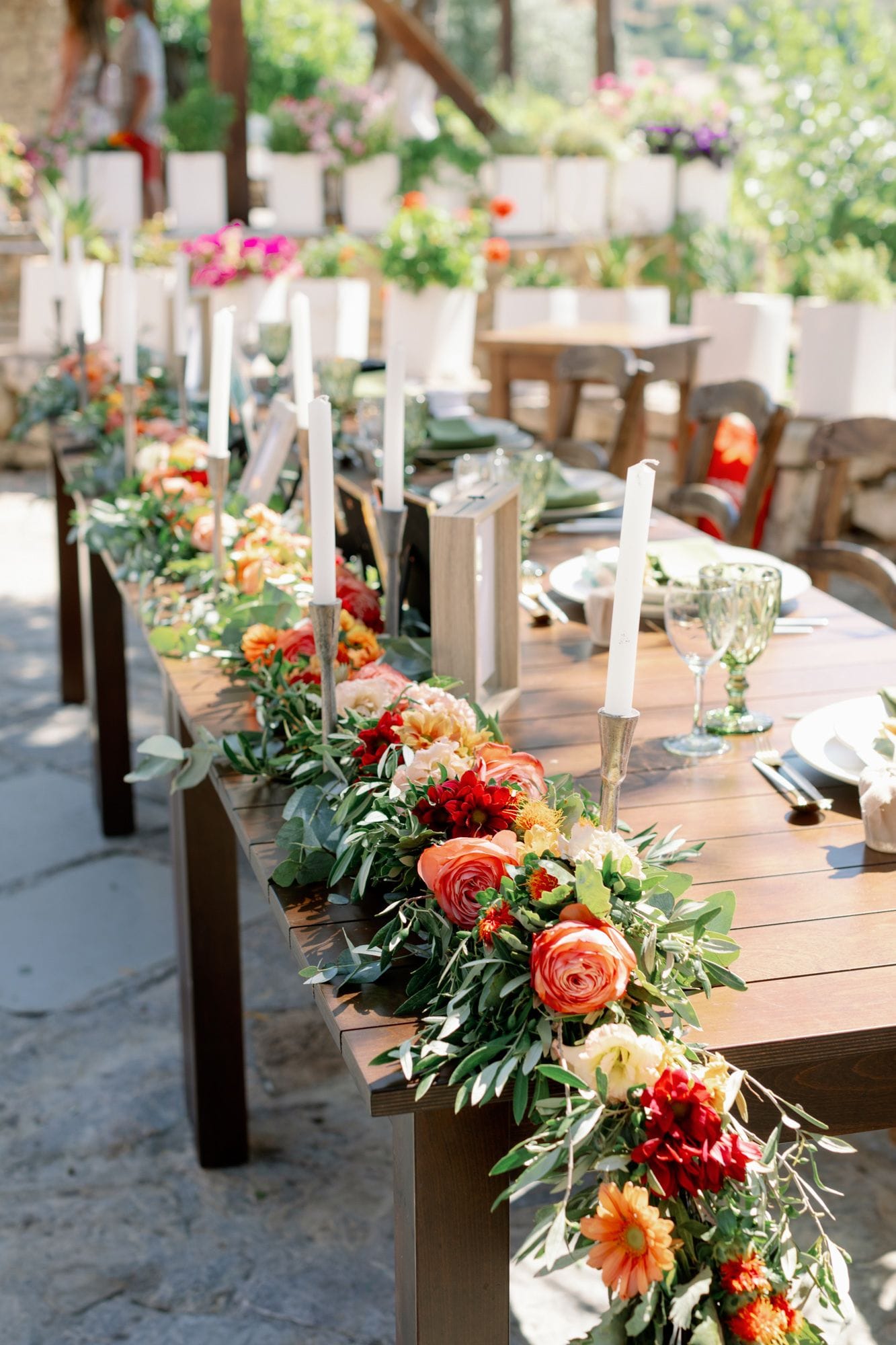 A long wooden wedding table runs diagonally across an outdoor terrace at Agreco Farm in Greece, dressed with a lush runner of coral and orange garden roses, dahlias, cream blooms and trailing olive and eucalyptus foliage. Tall white taper candles rise from silver candlestick holders between the blooms, and place settings with green-tinted glassware and white plates sit along the table. Soft, warm sunlight and a blurred background of wooden chairs give the scene a relaxed, golden-hour feel.