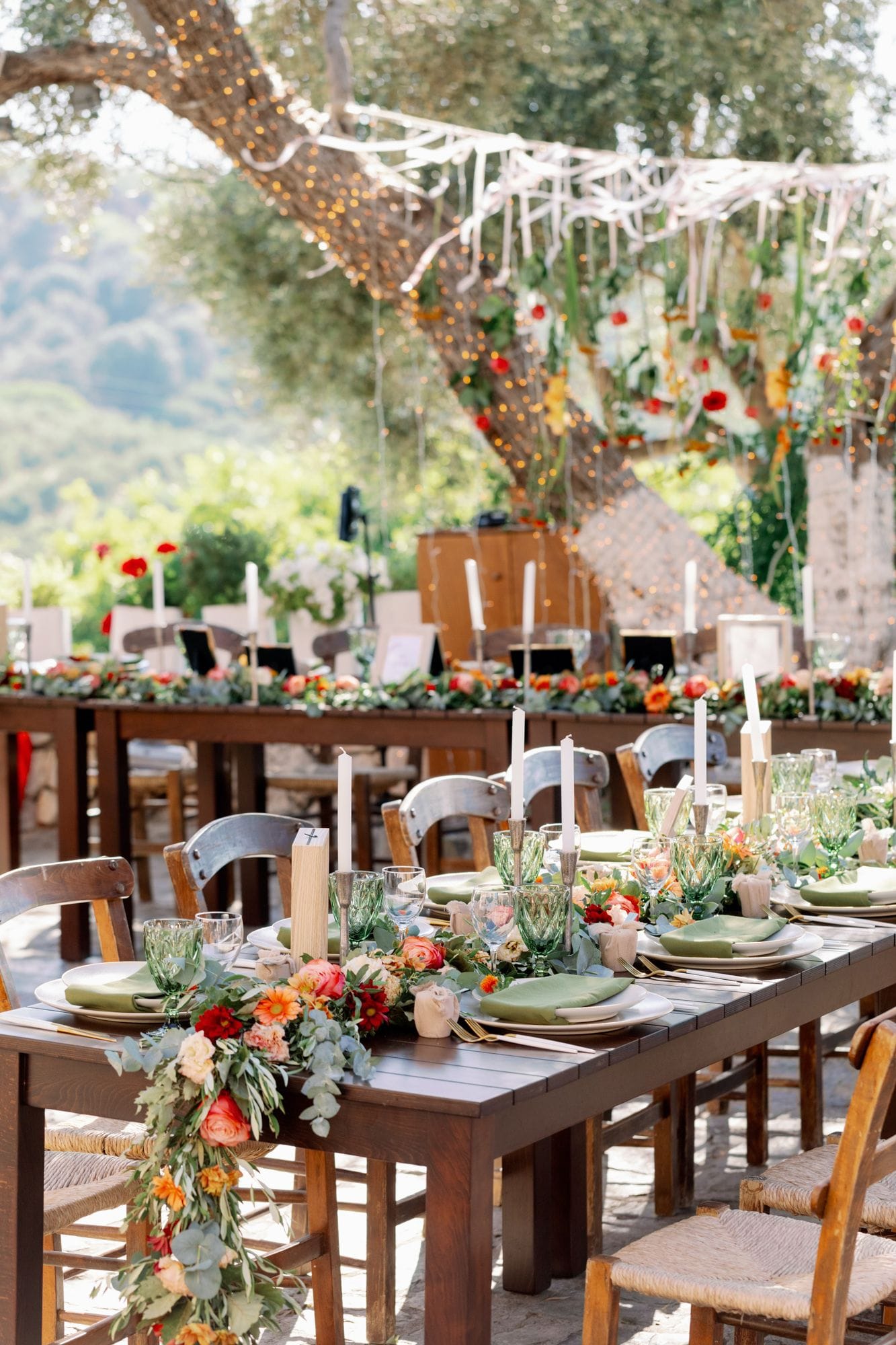 Long wooden farm tables under dappled sunlight are set for a wedding dinner at Agreco Farm in Greece, with trailing garlands of eucalyptus, olive leaves and orange and coral blooms, including garden roses, gerbera and small spray roses, running down the centre. Green-tinted glasses, sage-green linen napkins, white tapered candles and rustic wooden place cards complete each setting, while chairs with woven seats and tall candles line the way toward a softly lit, leafy courtyard.