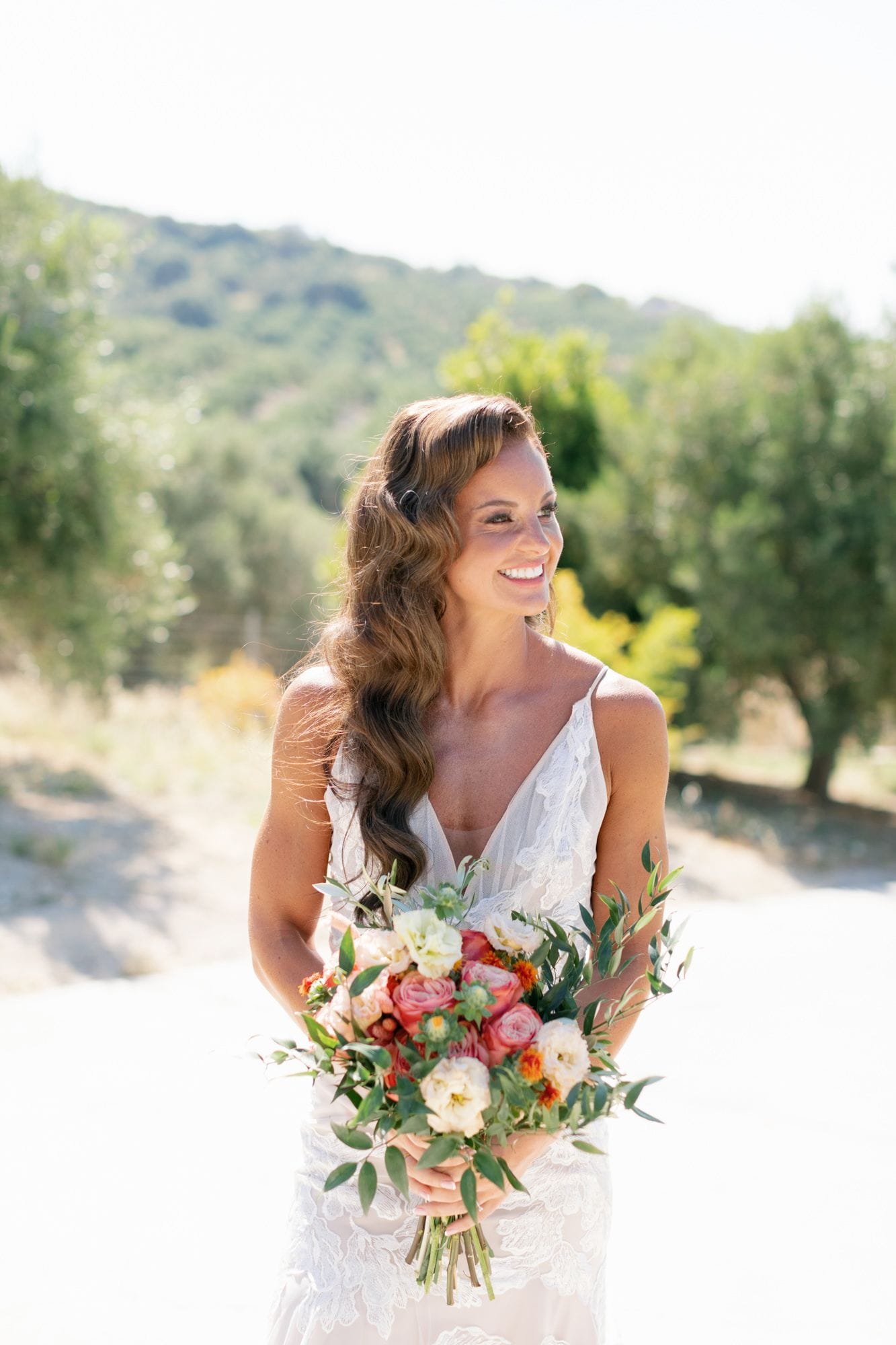 A smiling bride with long, softly waved hair falling over one shoulder stands on a sunlit path, holding a bridal bouquet of pink garden roses, cream lisianthus, small orange marigold-like blooms and trailing olive branches. Her white lace slip dress has thin straps and a V neckline, and olive trees blur softly behind her in warm light. This wedding bouquet was created by manfler for a celebration at Agreco Farm in Greece.