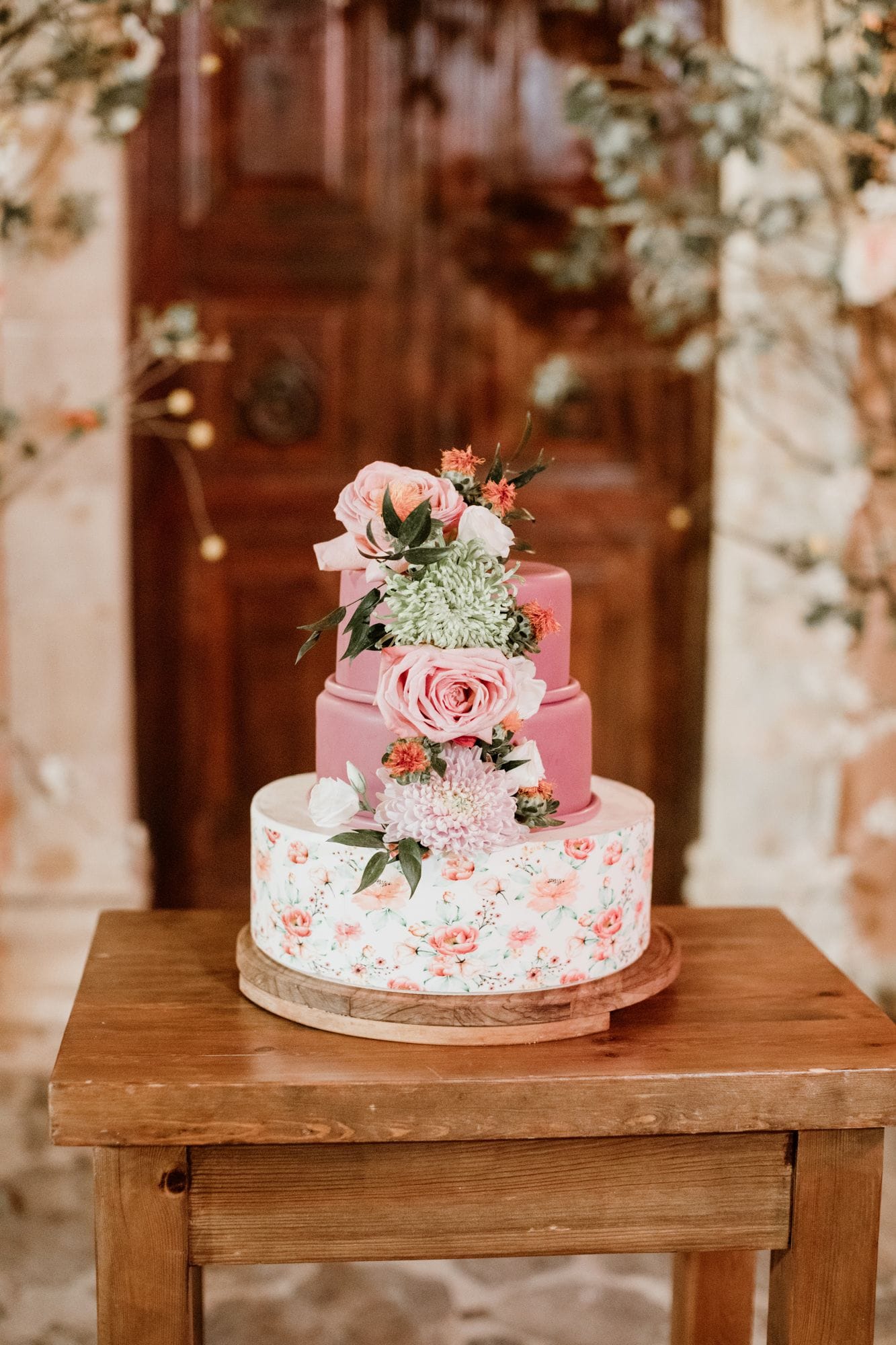 A three-tiered wedding cake stands on a wooden table in front of a large dark wooden door, with blurred floral garlands framing the scene. The upper tiers are dusty pink, and the bottom tier is white with a delicate pattern of pink and peach blooms. Pink garden roses, a pale green chrysanthemum, lilac blooms, orange accents and sprigs of dark green leaves cascade down one side, creating a soft, romantic wedding cake at Agreco Farm in Greece.