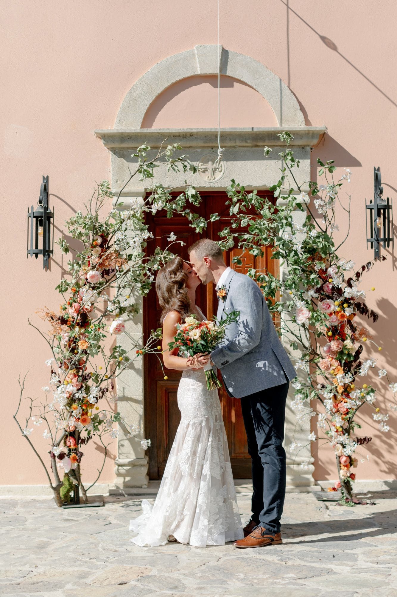 A bride in a lace fitted gown and a groom in a grey linen jacket share a kiss beneath a tall, arching frame of branches, white blossom, peach garden roses and orange and yellow blooms, with a bridal bouquet of coral and cream flowers in her hands. Behind them stands a dark wooden door set in a soft peach stone wall, lit by warm sunshine at Agreco Farm in Greece, where this wedding floral design was created.