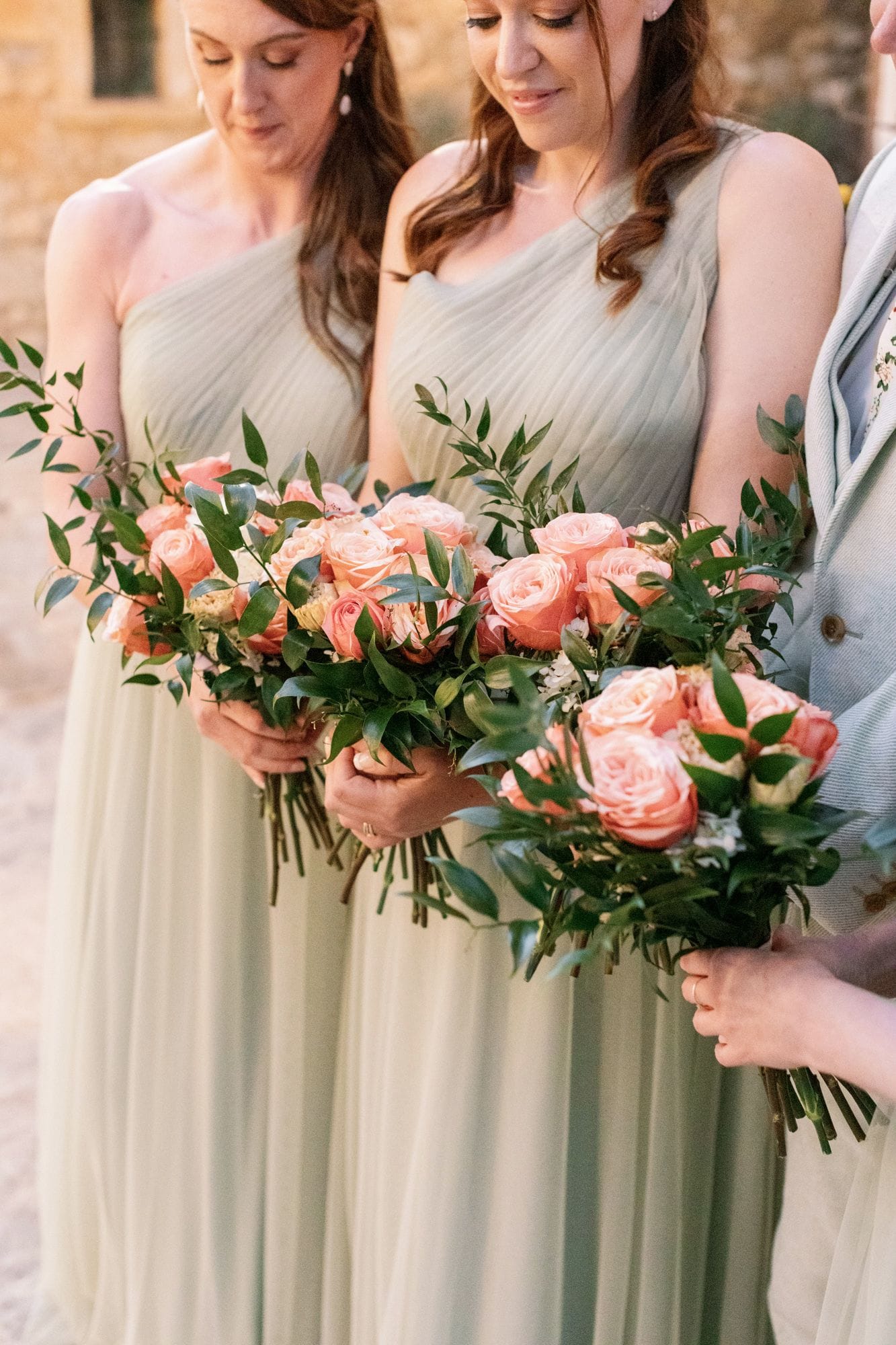 Three bridesmaids stand close together holding loose, rounded bouquets of peach and blush garden roses, with small white blooms and trailing sprigs of green foliage tucked among them. Their pale sage one-shoulder gowns have soft pleated fabric, and the warm, gentle light suggests a late afternoon. These wedding bouquets were arranged for a celebration at Agreco Farm in Greece.