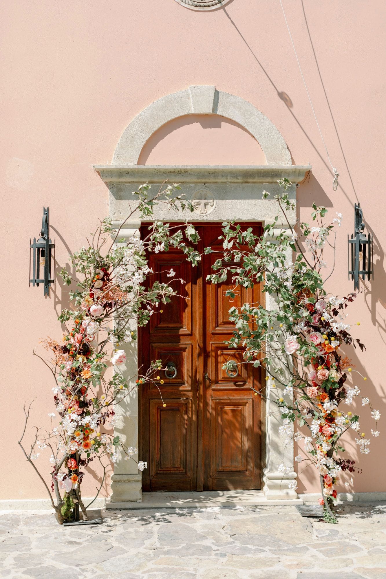 Two tall wooden doors in a pale stone frame are wrapped on both sides with floral garlands of white blossom, soft pink garden roses, and orange and apricot blooms, threaded through green branches with yellow craspedia balls scattered throughout. This wedding flower installation at Agreco Farm in Greece frames the entrance against a blush-pink wall, with dark iron lanterns mounted on either side in warm, soft daylight.
