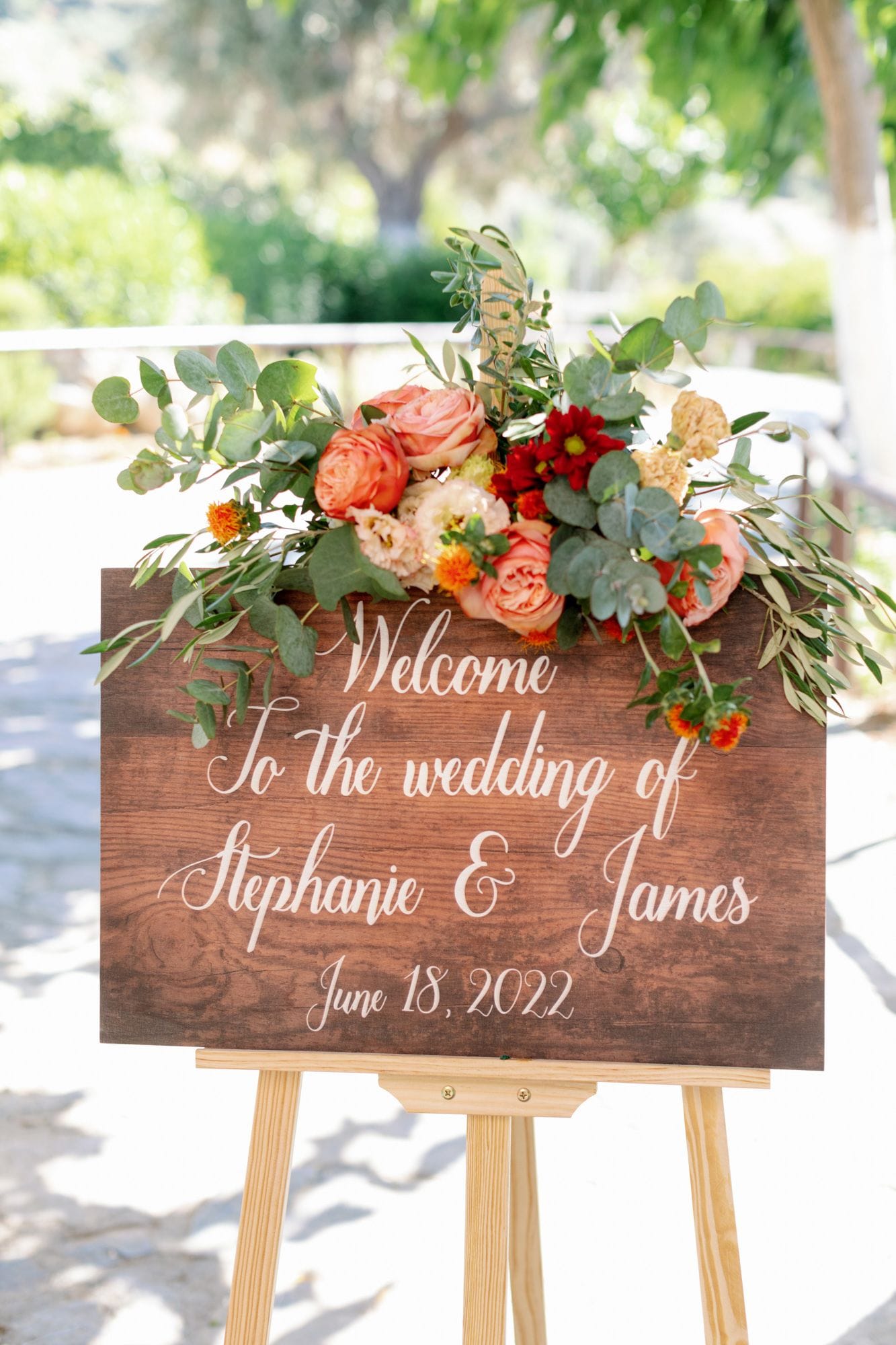 A wooden welcome sign with white script reading "Welcome to the wedding of Stephanie & James" is crowned with a lush arrangement of coral garden roses, lisianthus, burgundy and orange chrysanthemums, and apricot carnations, all tucked among silvery eucalyptus and trailing olive branches. The wedding flowers are set outdoors at Agreco Farm in Greece, with soft, sunlit greenery blurred behind them.