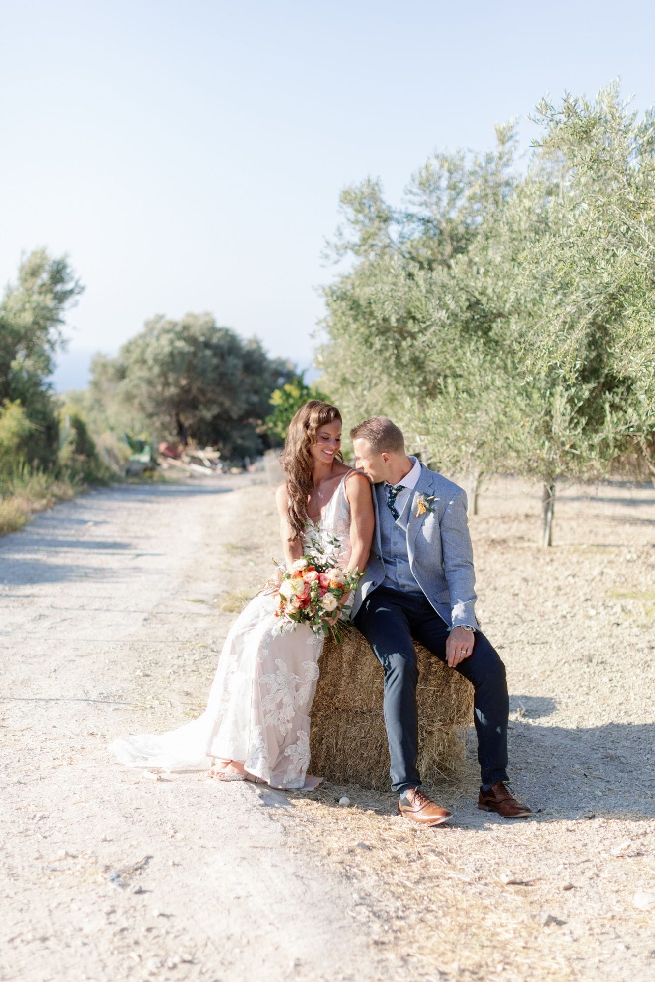 A bride in a blush lace gown and a groom in a light blue textured suit sit together on a round hay bale beside a gravel path, lined with silvery olive trees at Agreco Farm in Greece. The bride holds a wedding bouquet of coral and cream garden roses with eucalyptus, and she smiles as the groom leans in close to her shoulder. Soft, warm sunlight filters through the trees, giving the scene a relaxed, rustic and romantic mood.