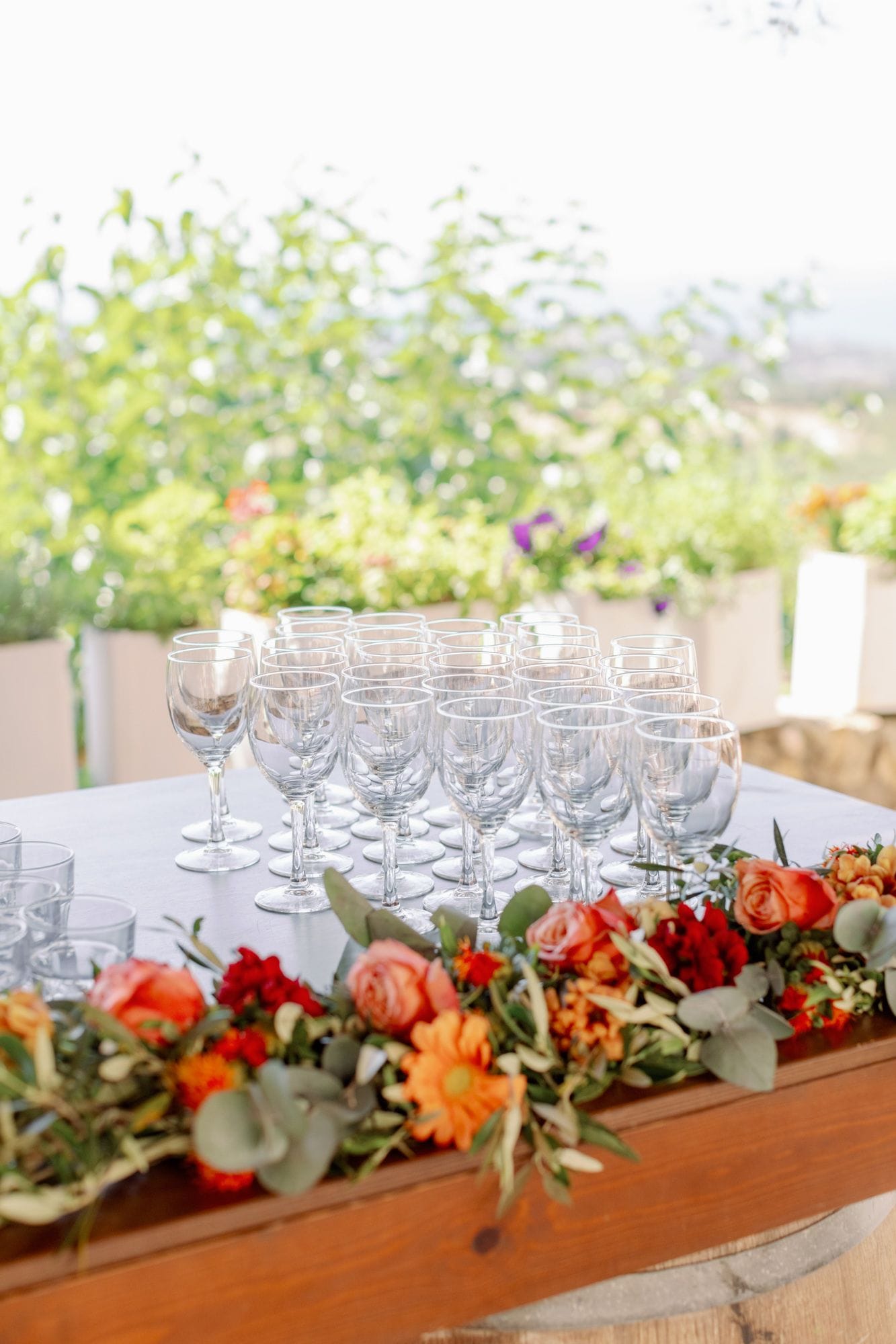 A long wooden trough runs across the foreground, filled with a garland of orange garden roses, red carnations, orange gerberas and small yellow blooms, woven through with silvery eucalyptus and olive sprigs. Behind it stands a dense rank of clear stemmed wine glasses on a white tablecloth, catching soft daylight. This wedding table flower arrangement was created by manfler for Stephanie and James at Agreco Farm in Greece, with warm autumnal tones.