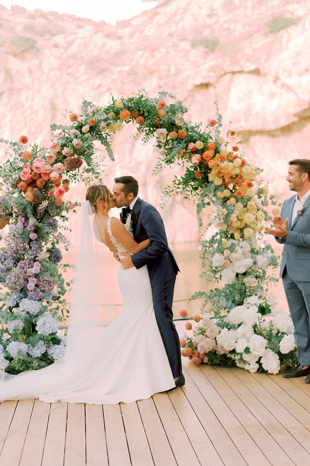 A couple shares a kiss beneath a sweeping wedding arch at Lake Vouliagmeni in Greece, framed by flowers that fade from blush pink roses and dusky lilac hydrangeas on the left to burnt orange roses and soft yellow blooms on the right. Trailing eucalyptus and greenery wind through the curve, and white roses and hydrangeas fill the base. The bride, in a fitted lace gown with a long veil, leans in while the groom in a navy suit holds her close, and a smiling man in a light blue suit claps beside them against a soft pink rocky backdrop.
