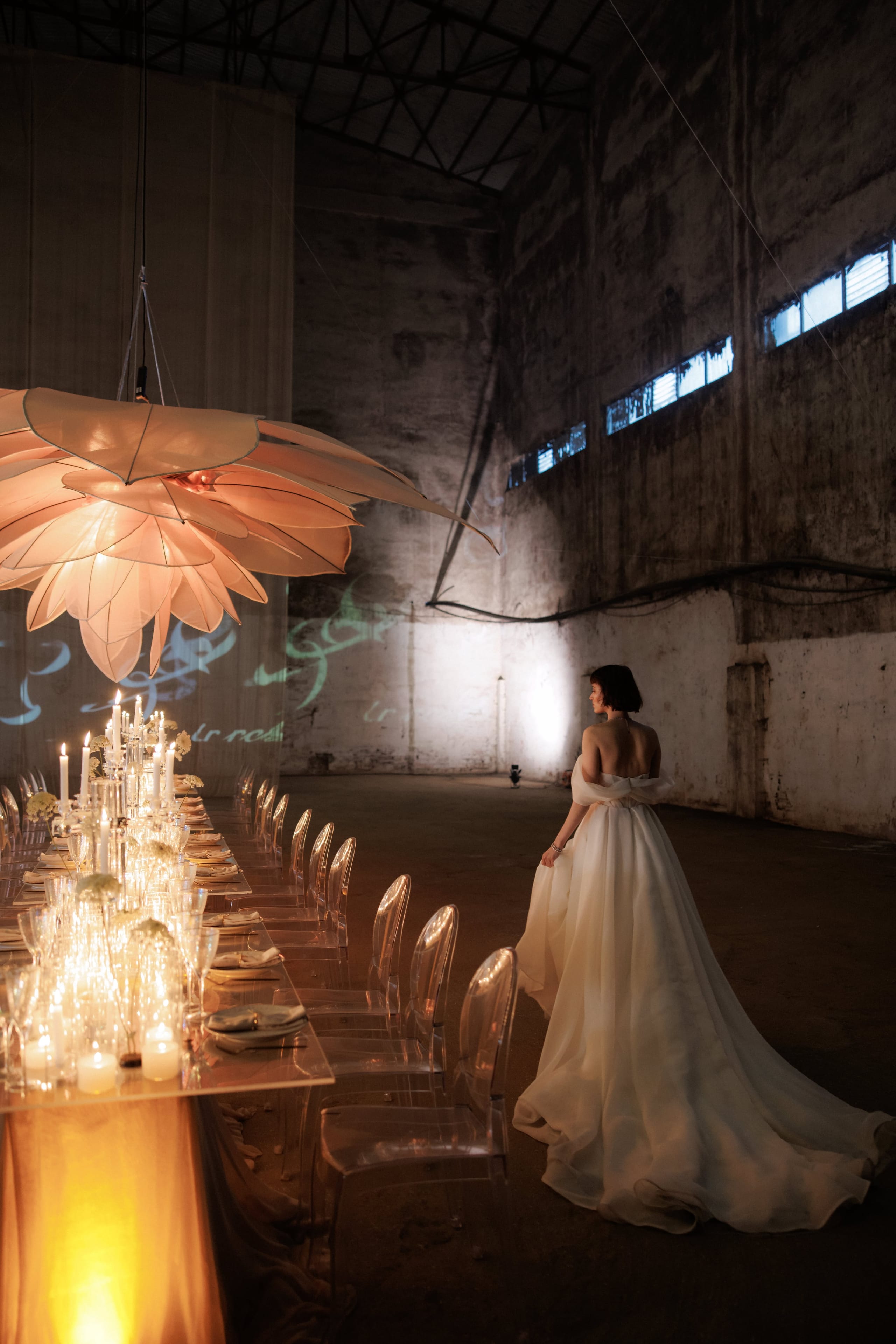 A bride with a short dark bob, seen from behind in an off-shoulder ivory gown with a long flowing train, gazes toward a rugged concrete hall lit by warm spotlights. On the left, a long table laid for a wedding dinner runs beneath a large glowing peach-coloured flower-shaped lantern, with clusters of white blooms and tall candles in glass vases and clear chairs. This wedding table setting is in Rethimno, Crete, Greece.