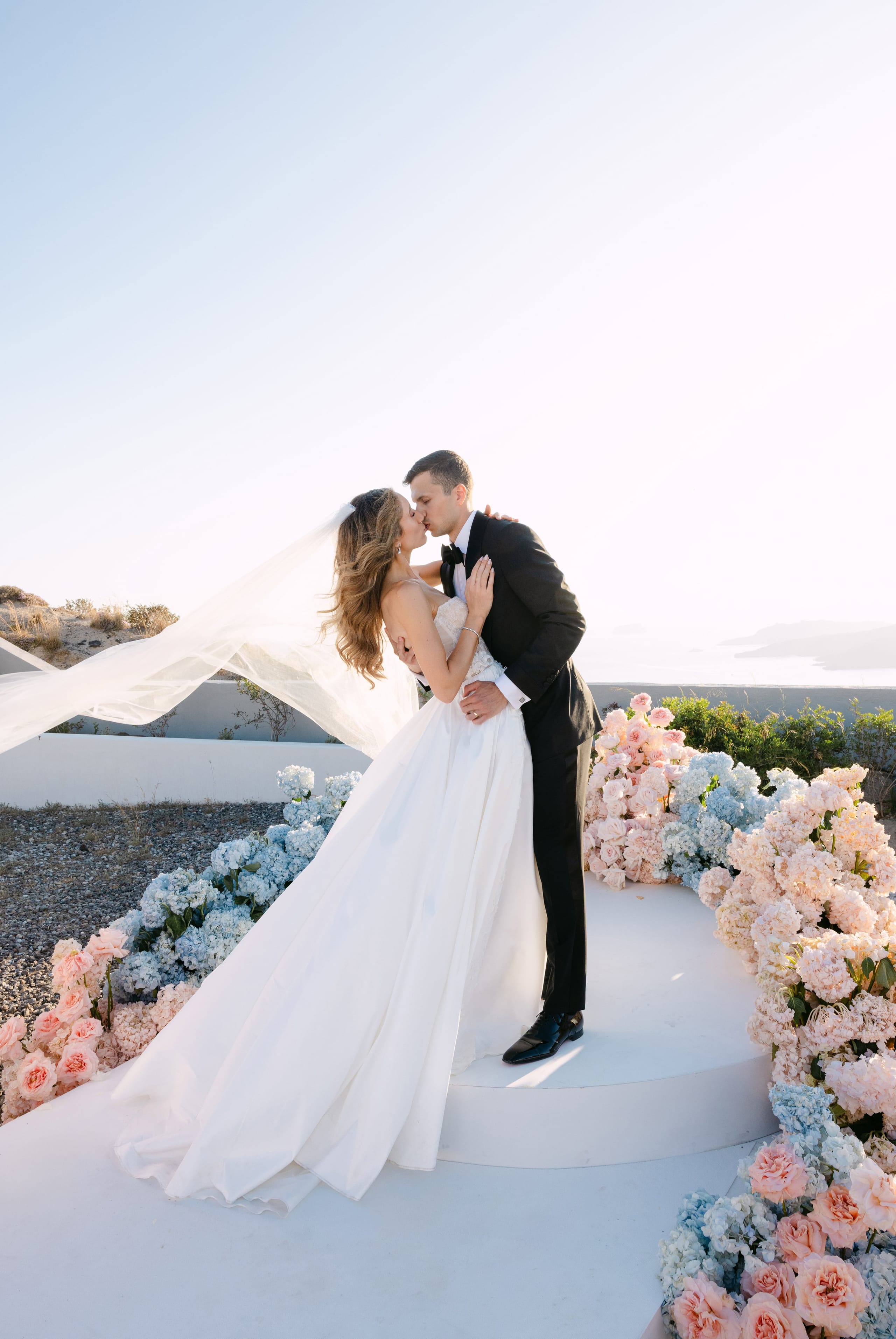 A bride and groom share a kiss on a whitewashed terrace in Santorini, Greece, at Eros Santorini, with a long veil streaming behind her in the breeze. She wears a strapless white satin gown and he a black tuxedo with a bow tie. Along the floor and edges of the terrace, wedding flowers in soft blue hydrangeas, blush garden roses and pale pink peonies spill in lush drifts toward a hazy sea and distant hills under a bright, pale sky.