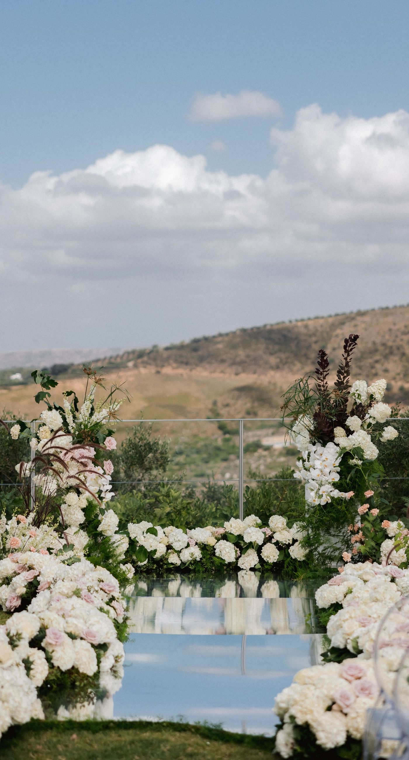  Sasha & Alex - Amidst a breathtaking landscape, this exquisite showcase captures the essence of romance and elegance. Soft hues of blush and ivory blend seamlessly with lush greenery, creating an ethereal garden-like setting. The mirrored aisle reflects the joyful union, adding a touch of glamor to the scene. Delicate florals, thoughtfully arranged, frame the moment, enhancing the dreamy, sophisticated atmosphere. This outdoor celebration perfectly balances natural beauty with refined artistry, embodying the timeless allure of love in a serene, picturesque locale. - Photo 3