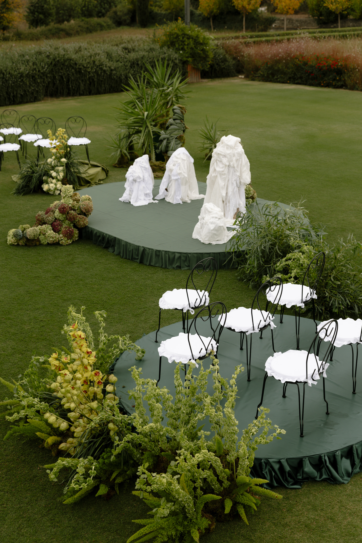 Seen from above, a lawn at Roxani Estate in Greece is set with a round green platform holding black wrought-iron bistro chairs, each with a ruffled white cushion, and a larger raised stage behind them where three tall, draped white forms stand among sculptural white fabric. Clusters of green-and-burgundy hydrangeas and yellow cymbidium orchids with pale green foliage frame the stage edges, with lush bamboo-like plants behind, all arranged as part of a wedding or event installation.
