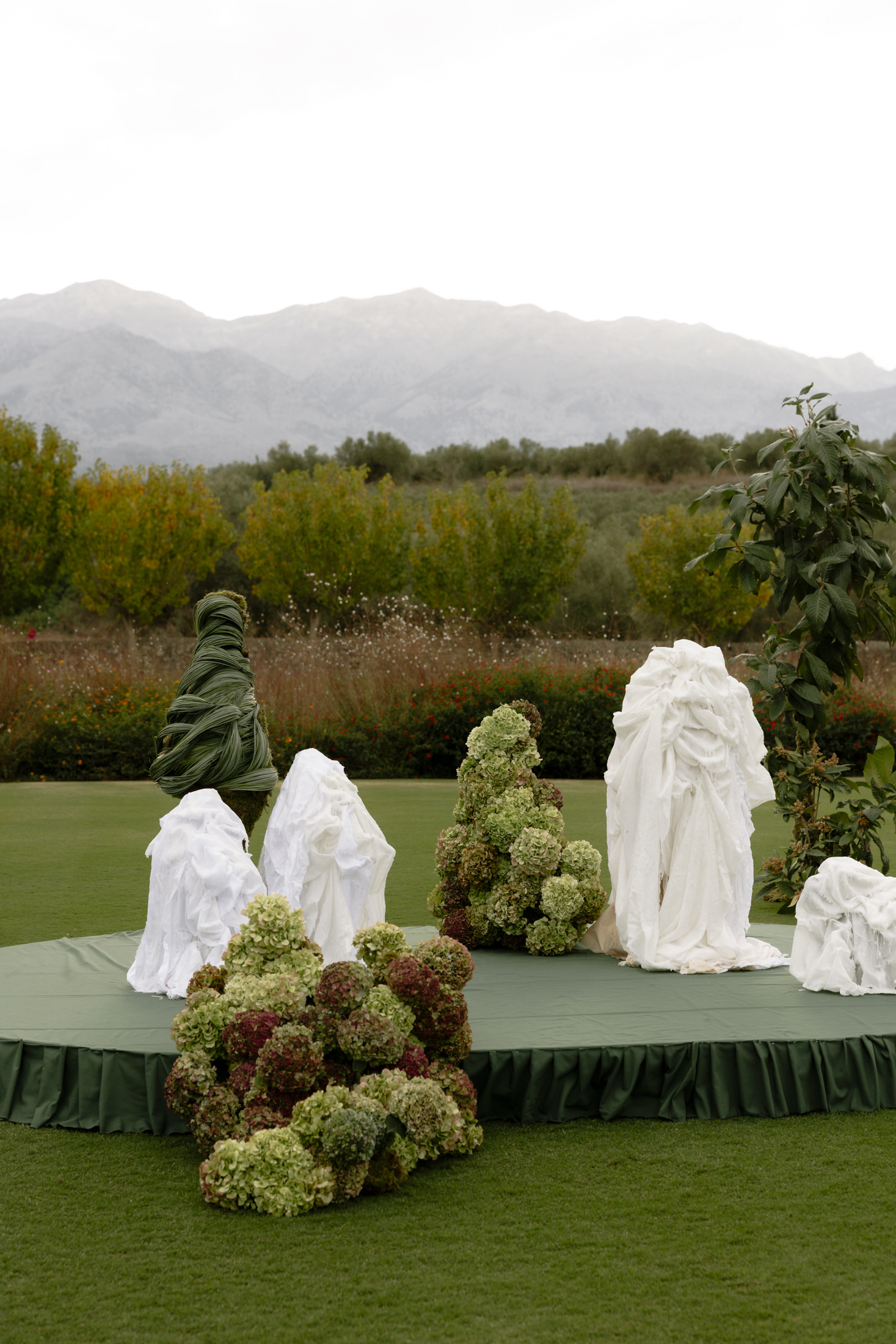 Sculptural installations on a green-skirted round platform stand on a lawn at Roxani Estate in Greece, part of the Green Edit wedding flower display. Three shrub-like forms are built from clusters of green and blush-tinged hydrangea heads, while a tall column is wrapped in twisted green foliage and three draped forms are swathed in white, rumpled fabric. Behind them, a row of flowering shrubs and olive-toned trees frames the soft, overcast light.