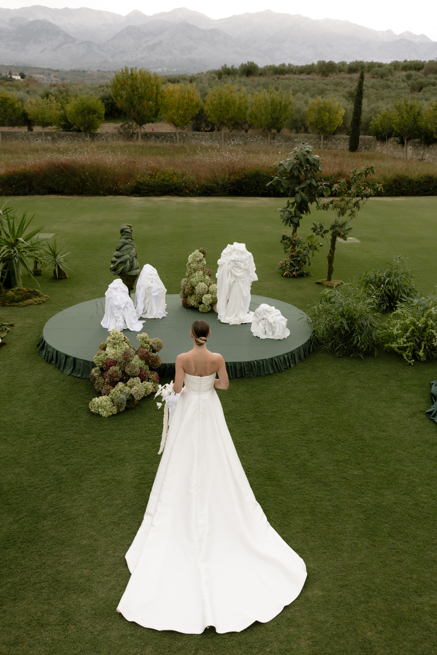 A bride in a strapless white satin gown with covered buttons down her back walks across a sweep of green lawn toward a round dark-green platform. On it stand tall sculptural arrangements of white draped fabric and clusters of green-and-burgundy hydrangeas, with a spiky green foliage piece and two young trees beside them. This wedding flower installation at Roxani Estate in Greece has a calm, garden-like mood, shot from above.