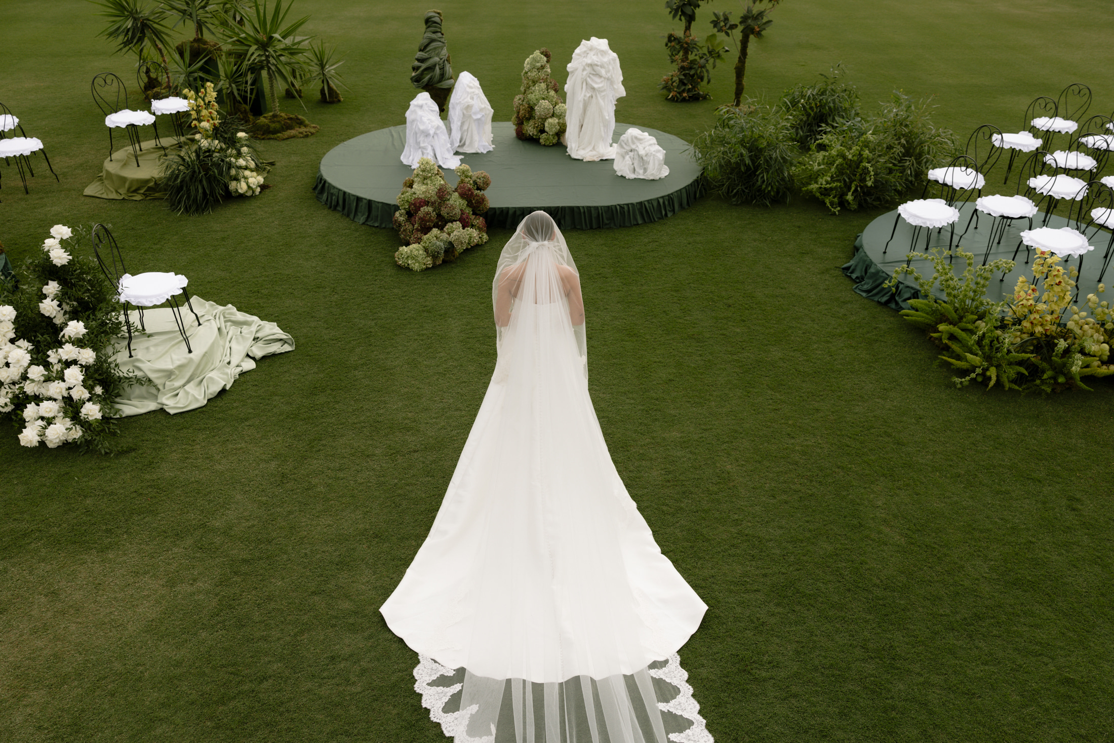 A bride seen from behind, in a satin gown and a long veil with lace edging that trails across a lawn, walks toward a round green-skirted platform of white draped forms and hydrangea clusters at Roxani Estate in Greece. Smaller white-topped chairs with green draped cloths and white rose arrangements line the grass, and the scene is a soft, muted green and ivory, shot from above. These are wedding flowers and ceremony styling.