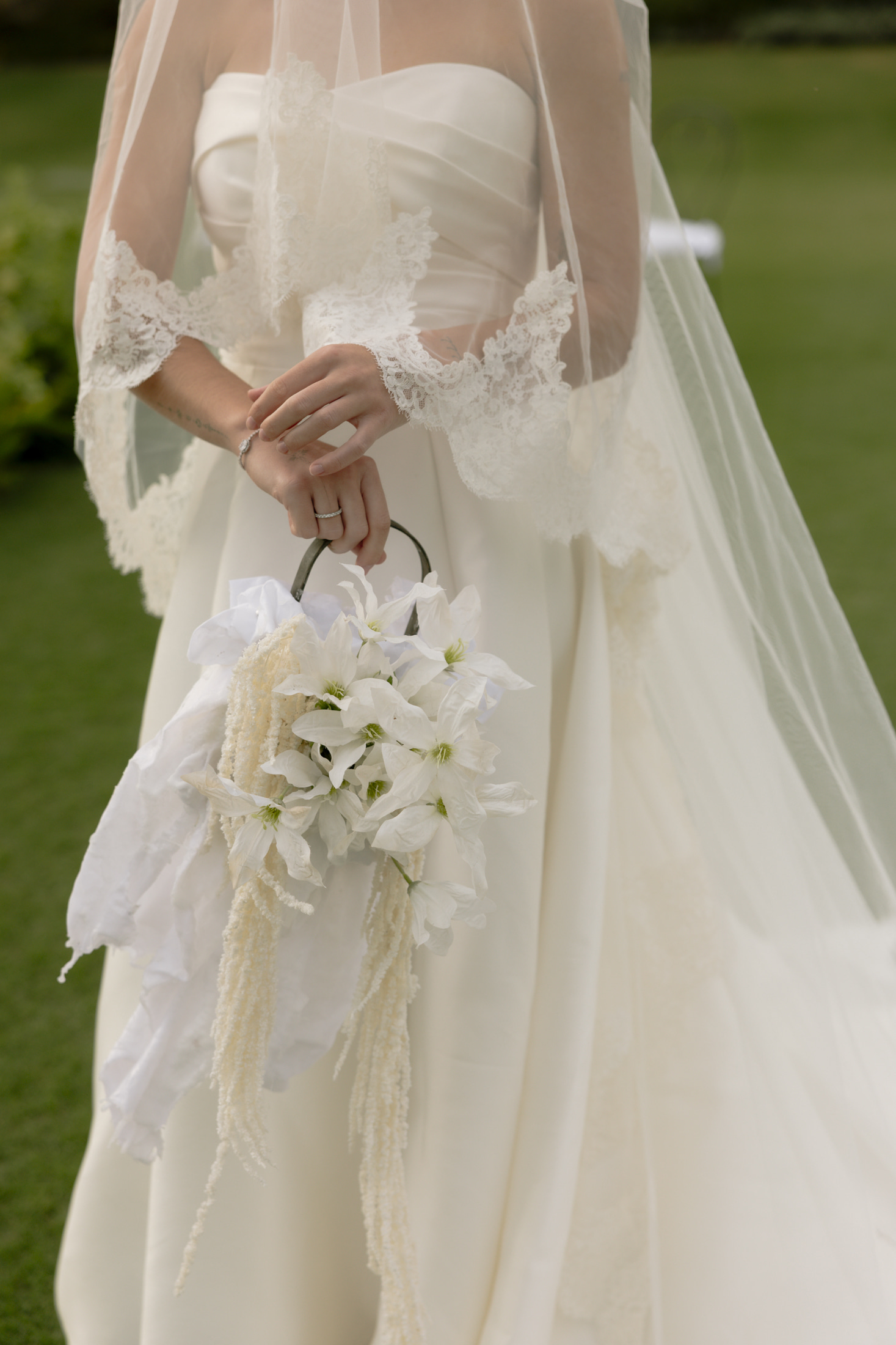 A bride in an ivory satin gown with lace-trimmed sheer sleeves and a long veil holds a white bouquet by a round metal handle, her hands clasped at her waist. The bouquet is made of white star-shaped blooms, possibly clematis, with cream fluffy foliage and white ribbon tied at the stems. Soft green lawn sets the scene at Roxani Estate in Greece, where this wedding flower design was created.