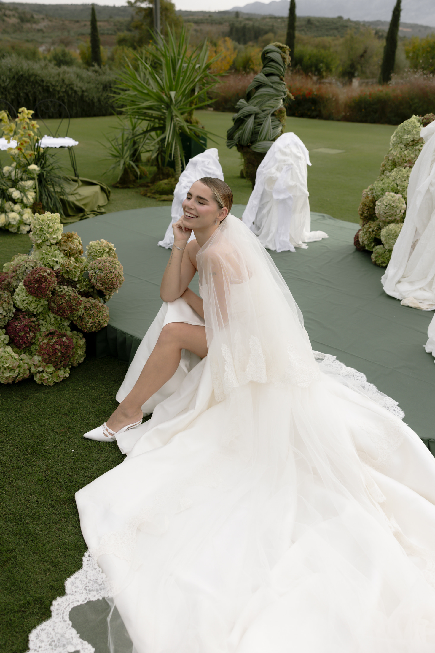 A bride in a strapless white gown and a sheer veil with lace edging sits on the edge of a round green platform, laughing with her hand under her chin. Behind her, shrouded white draped shapes sit among tall, sculptured greenery, and large clusters of green and blush hydrangeas form a mound at the left and right edges. The lawn is lush and the setting is a garden at Roxani Estate, a wedding venue in Greece, with wedding flowers arranged as part of the outdoor styling.
