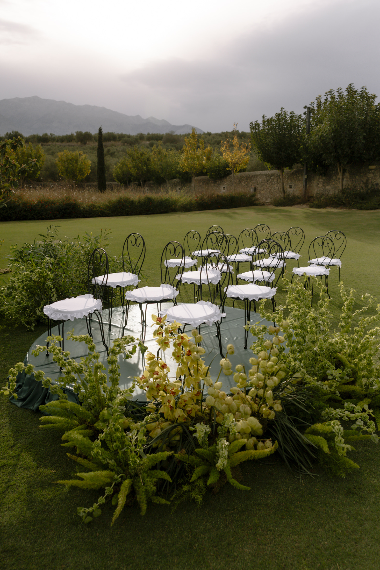 A circle of black wrought-iron bistro chairs with white ruffled seat covers sits on a pale green platform on a lawn, framed by lush green foliage and sprays of yellow cymbidium orchids and pale green bells-of-Ireland-like stems in the foreground. Behind, a low stone wall, clipped trees and olive groves stretch toward distant hills under soft daylight. This is a wedding ceremony set-up with floral styling at Roxani Estate in Greece.